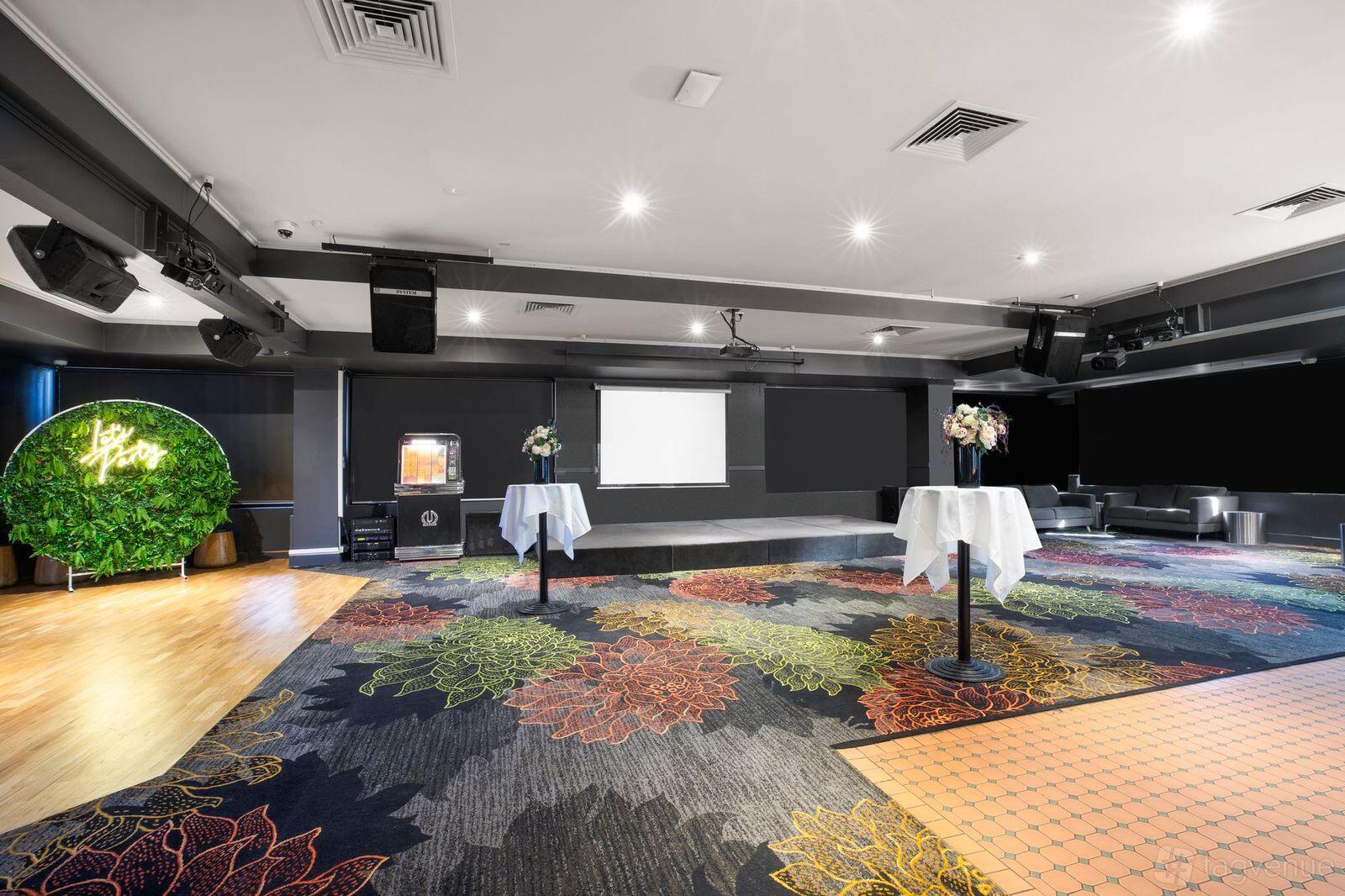 An event space in a pub with a floral-patterned carpet, stage, cocktail tables, and a neon sign with greenery at Burvale Hotel.