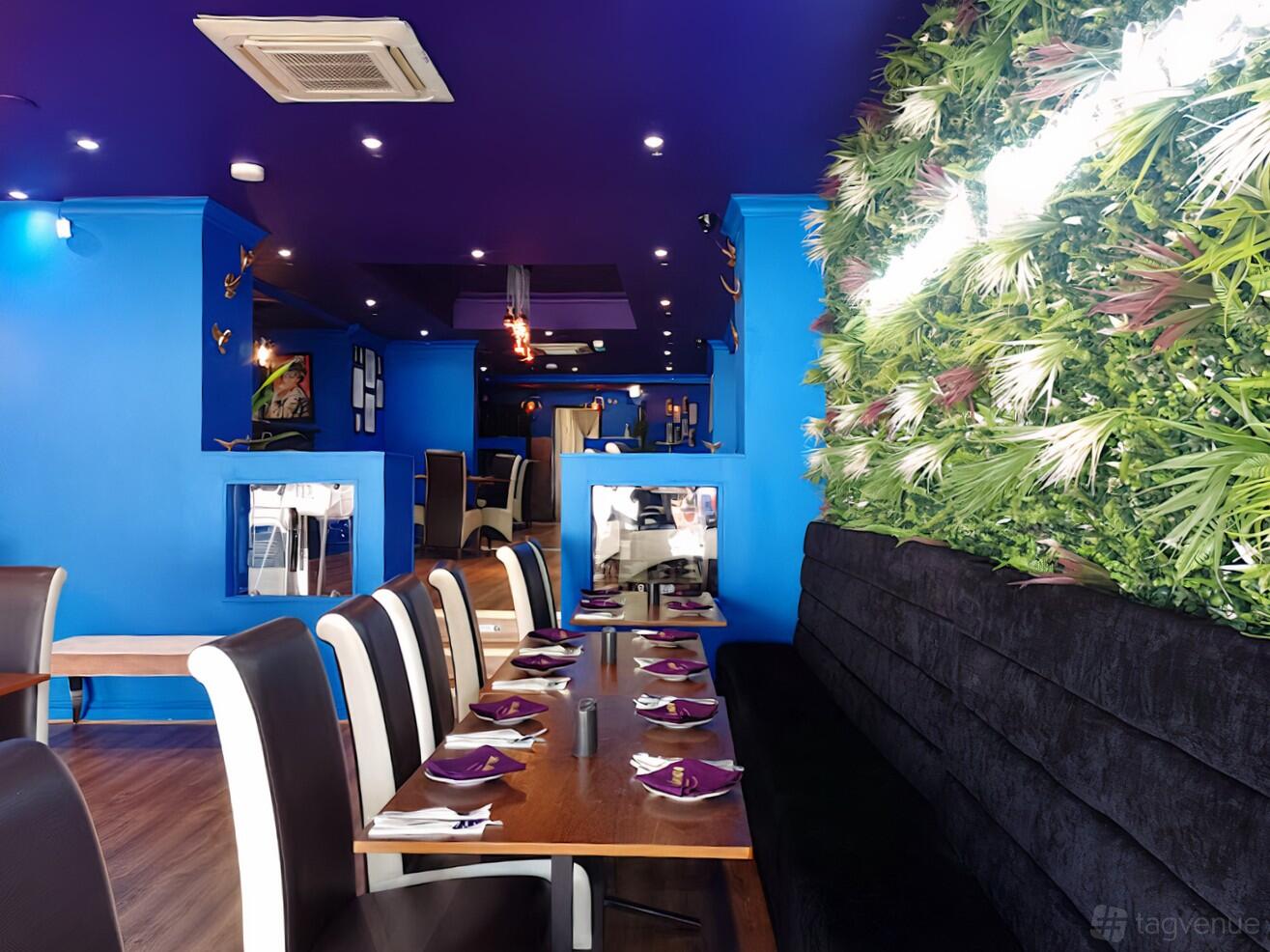 A restaurant with blue walls, long wooden tables set for dining, and a green plant feature wall at Aladdin Restaurant.