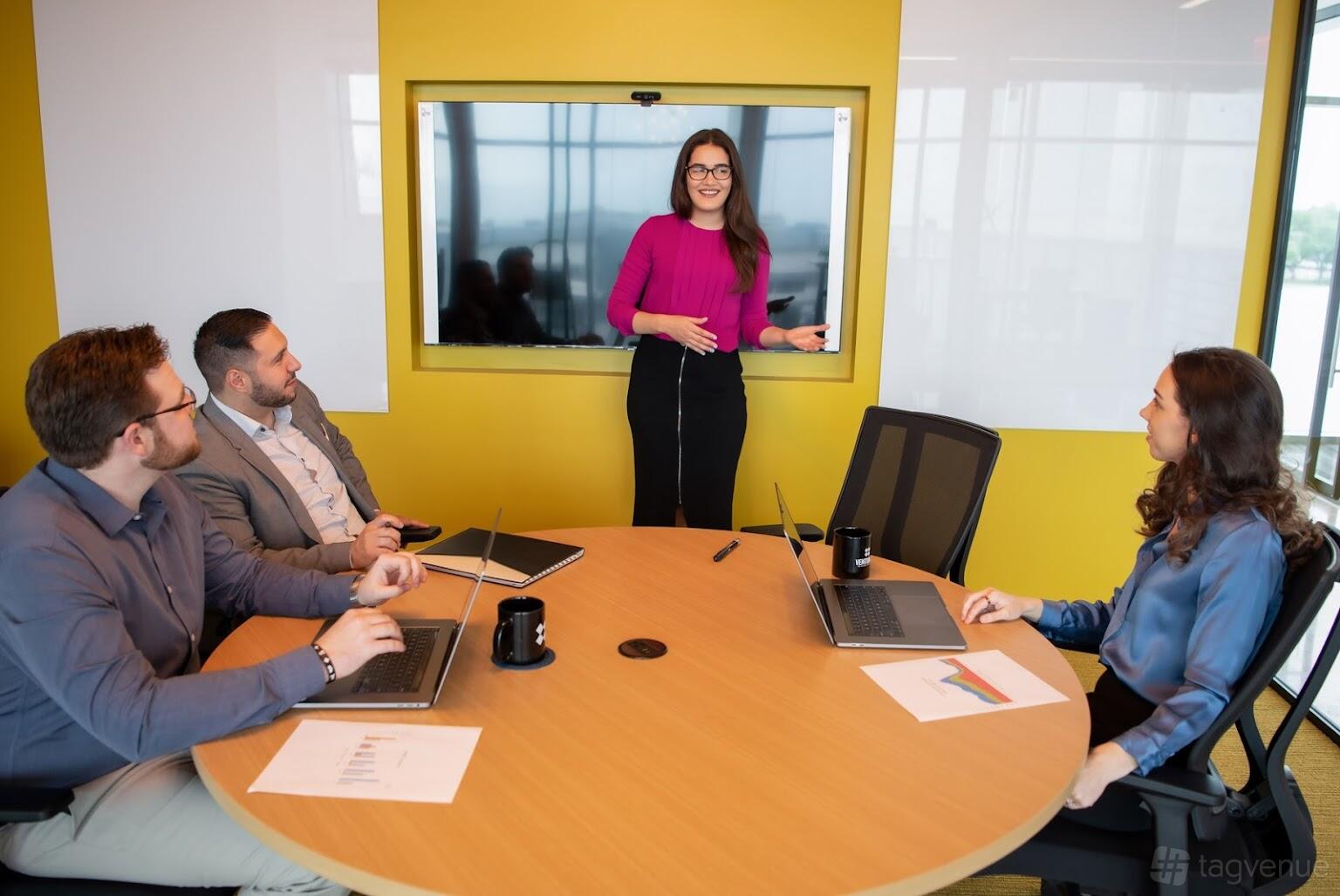 A coworking meeting room with a round table, four people, laptops, and a wall-mounted screen at Venture X Dallas – Braniff Centre.