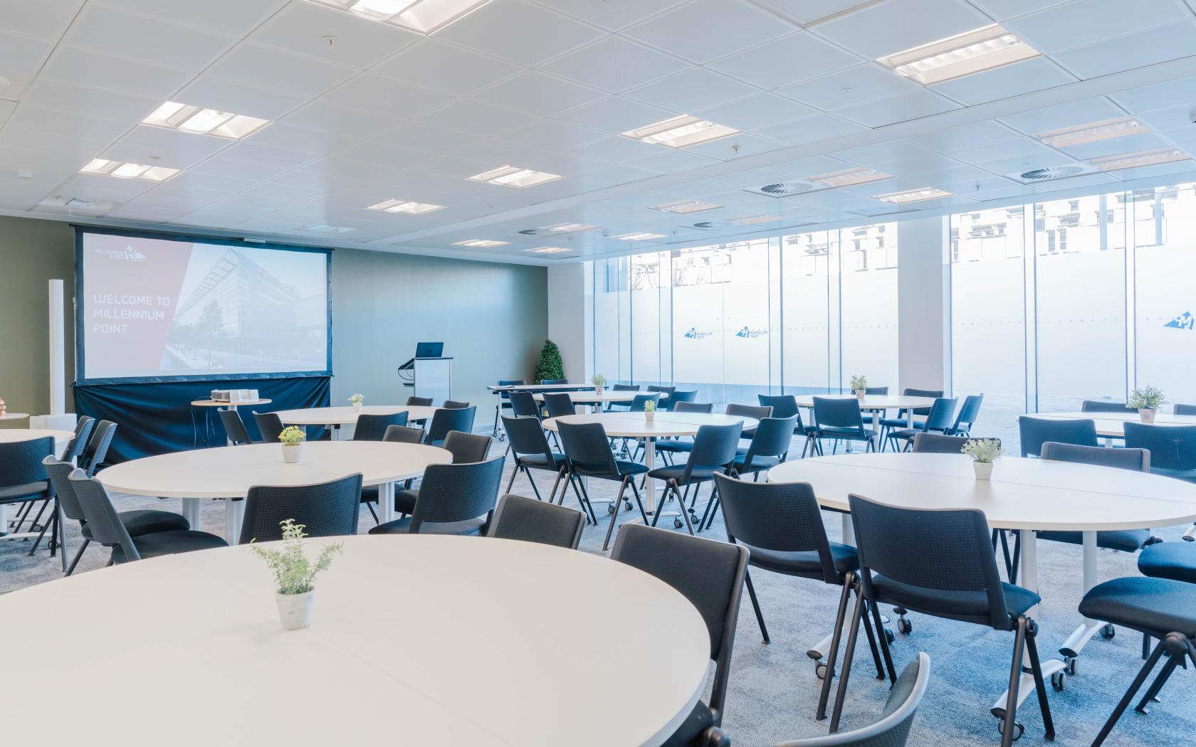 An event venue with round tables, black chairs, a projector screen, and floor-to-ceiling windows at Millennium Point.