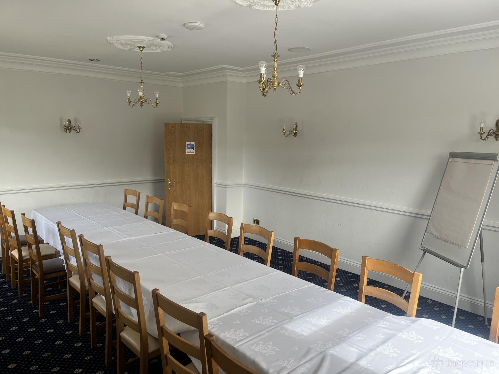 A function room with long white-clothed tables, wooden chairs, chandeliers, and a flip chart at The Manor Golf Club.