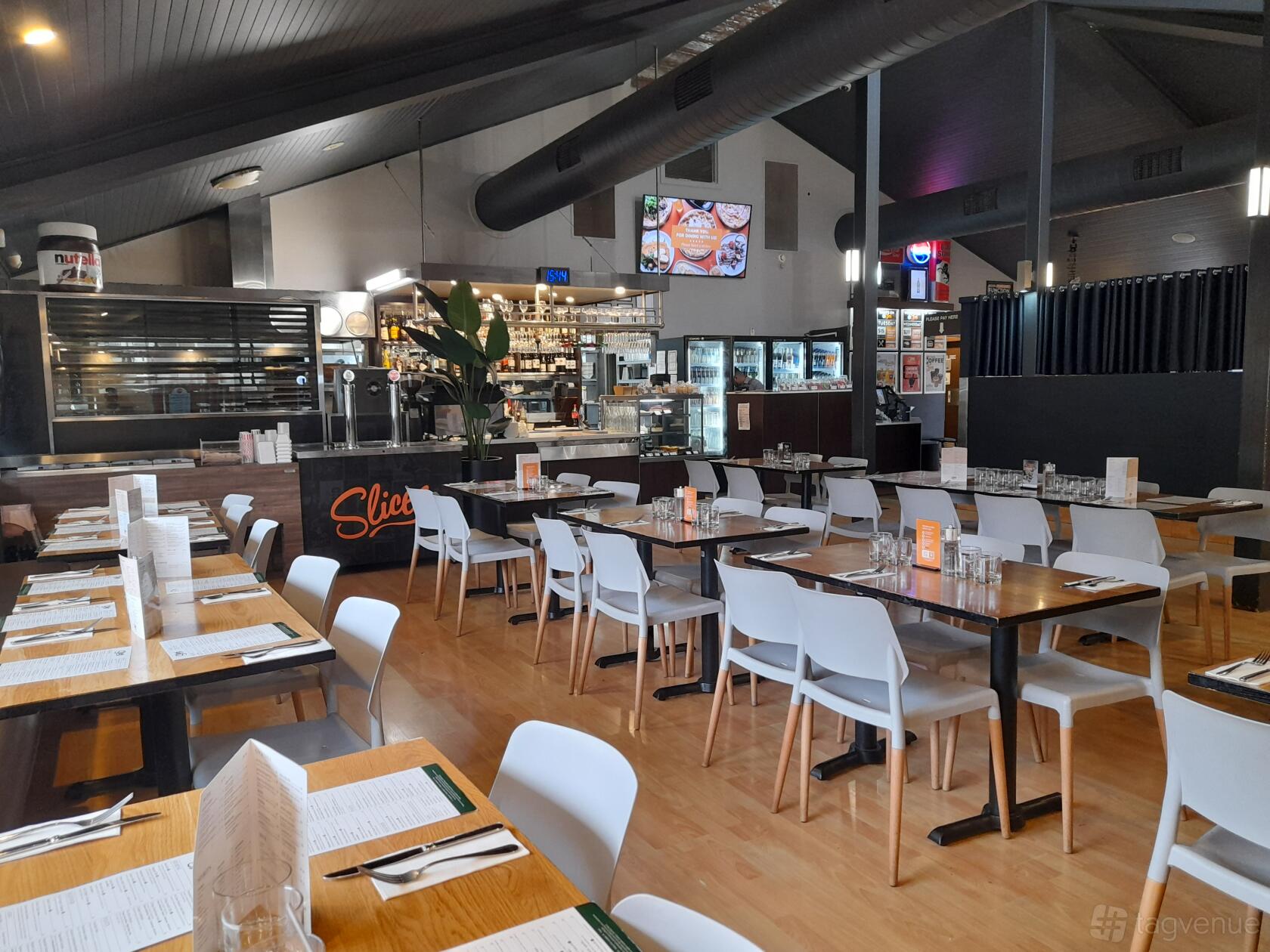 A restaurant with wood flooring, white chairs, set tables, and an open kitchen at Slices Keilor.
