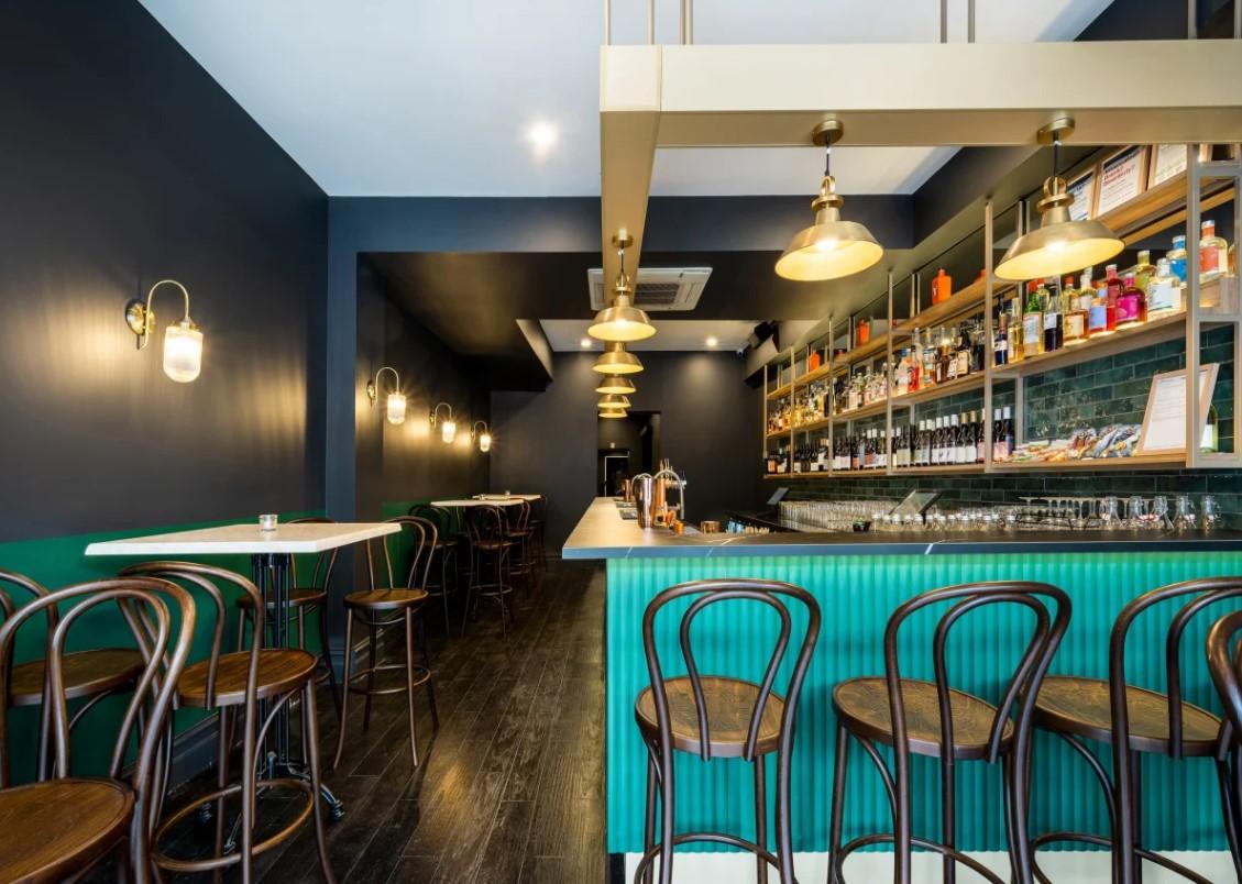 An event space with a long green bar, gold shelving, dark wood chairs, and wall-mounted lights at Blackwoods Wine and Cocktail Bar.