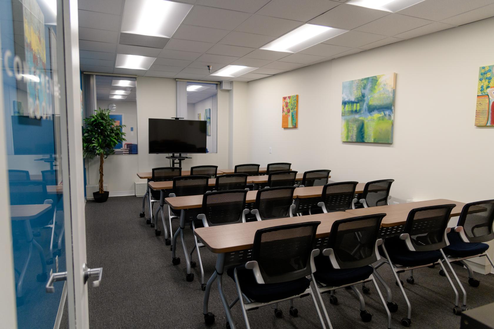 A meeting room with rows of tables and mesh chairs facing a large screen and potted plant at Creative Colony Spaces.