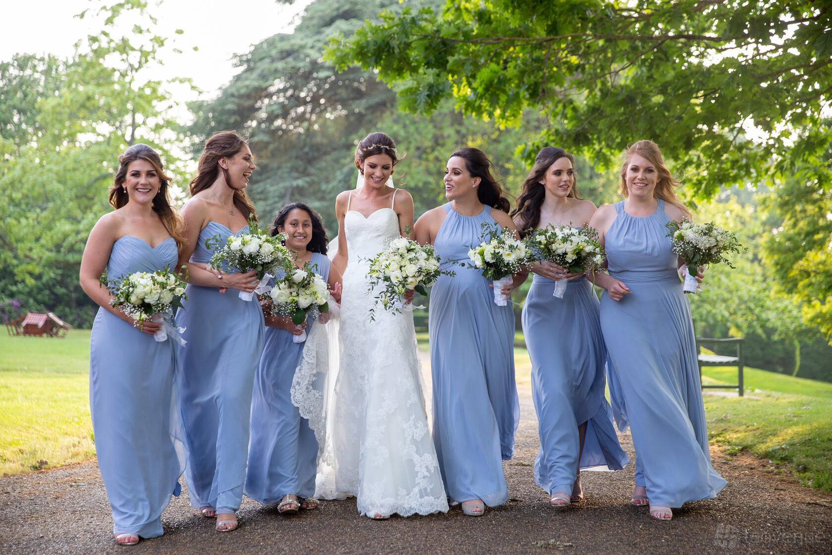 Bridesmaids in blue dresses walking with the bride along a tree-lined path at Oatlands Park Hotel grounds.