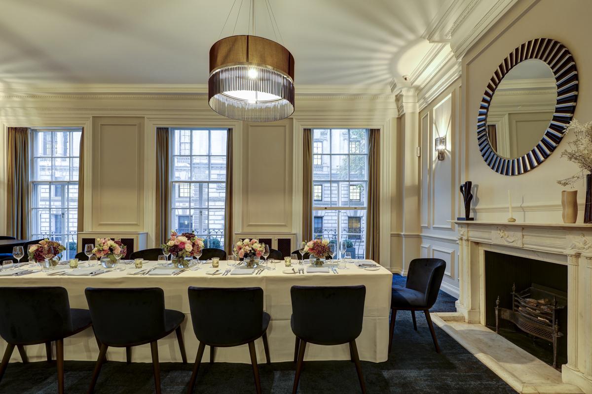 An event space with large windows, a long dining table set with floral centerpieces, and a decorative fireplace at Surveyors House at Rics Westminster.