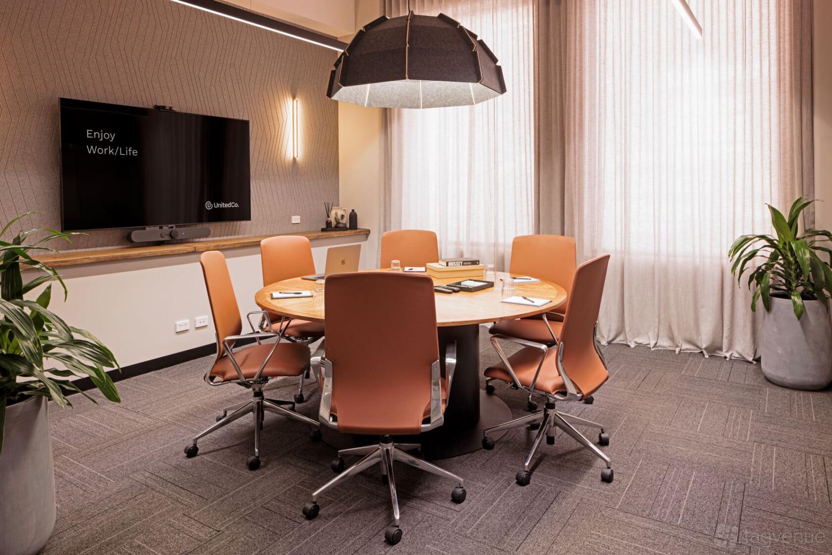 A meeting room with a round wooden table, six orange chairs, wall-mounted screen, and floor-to-ceiling curtains at United Co. Fitzroy.