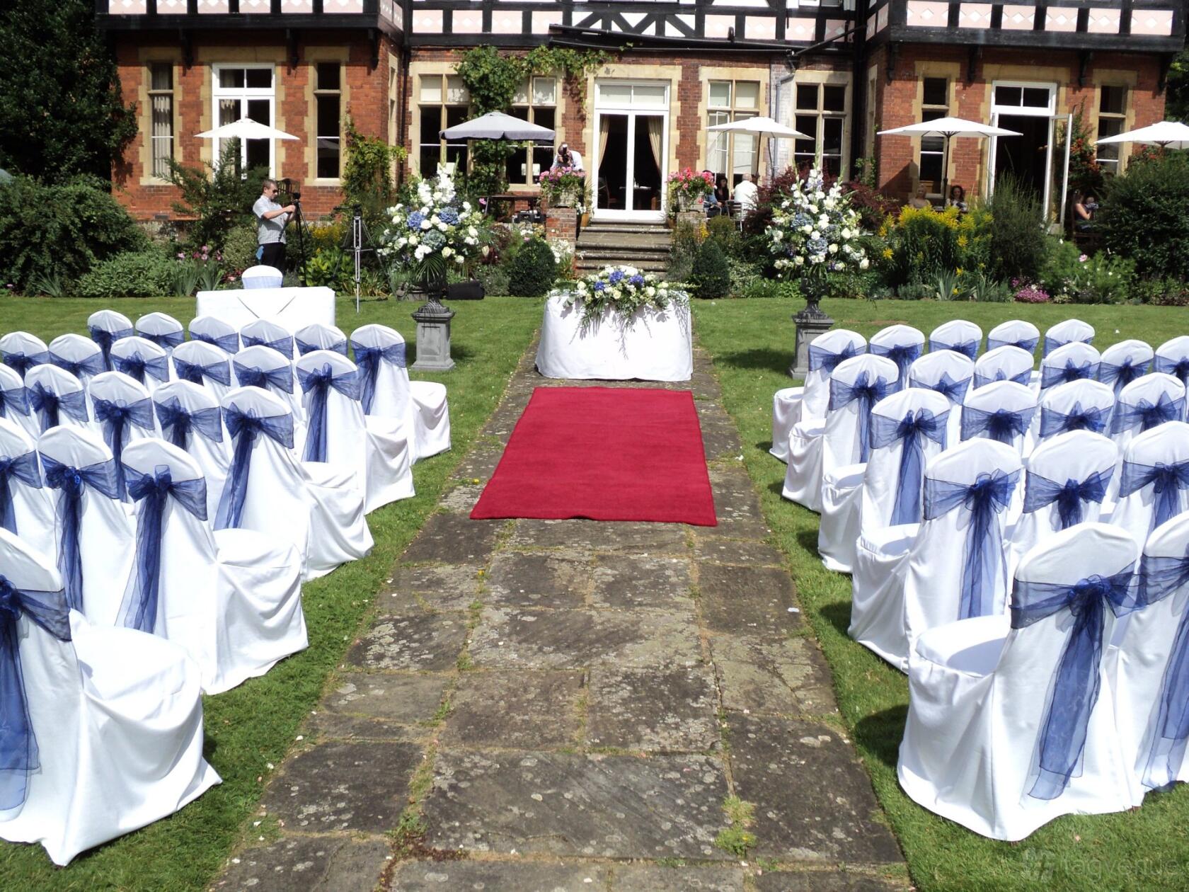 An outdoor event space with rows of white chairs tied with navy bows and a red aisle runner at The Grange Country House Hotel.