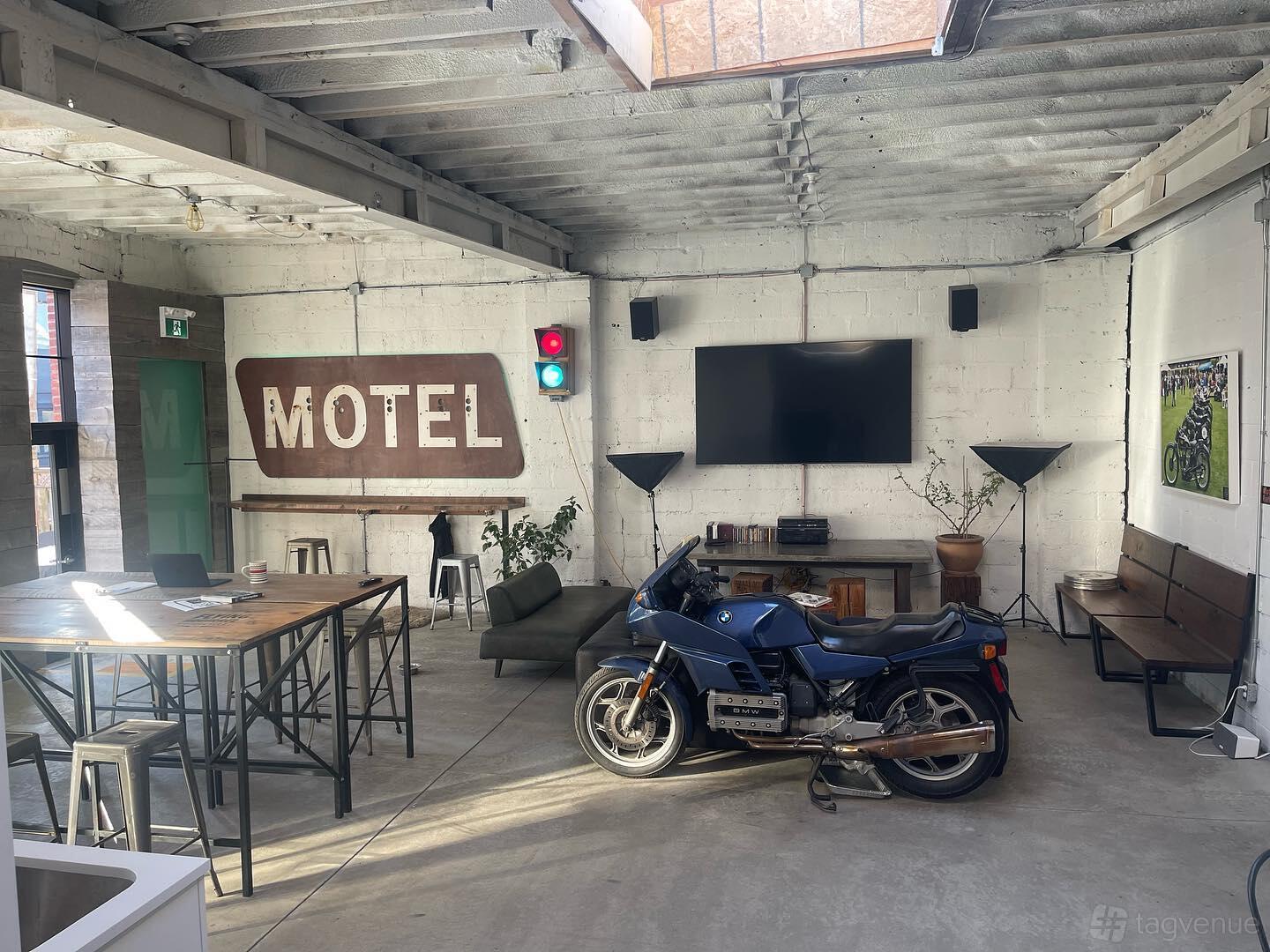 A cafe lounge with concrete floors, a motorcycle display, vintage motel sign, and wall-mounted TV at Flying Squirrel Motorcycle Co.