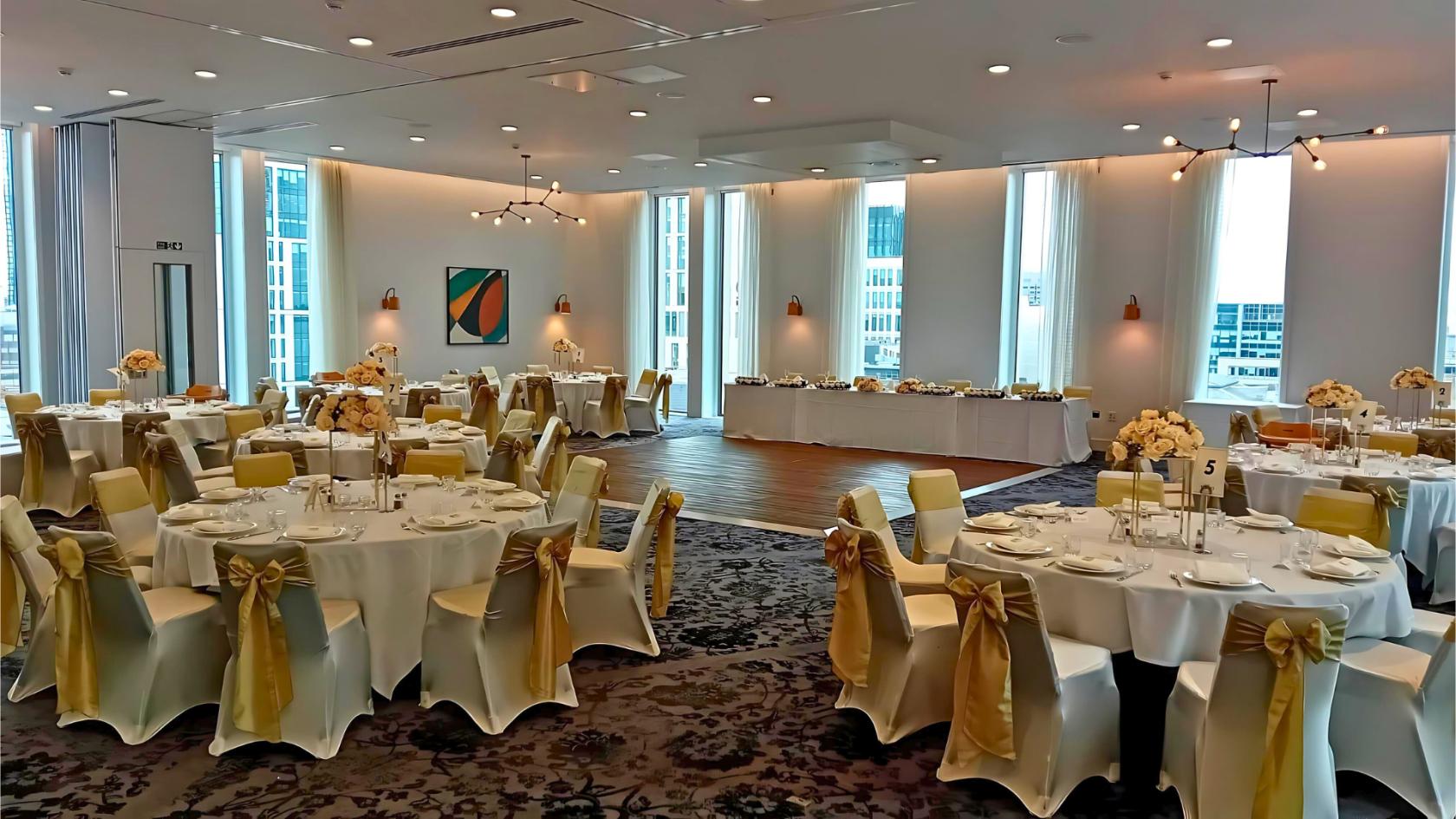 A banquet hall with floor-to-ceiling windows, round tables with white linens, and gold chair sashes at voco Manchester.