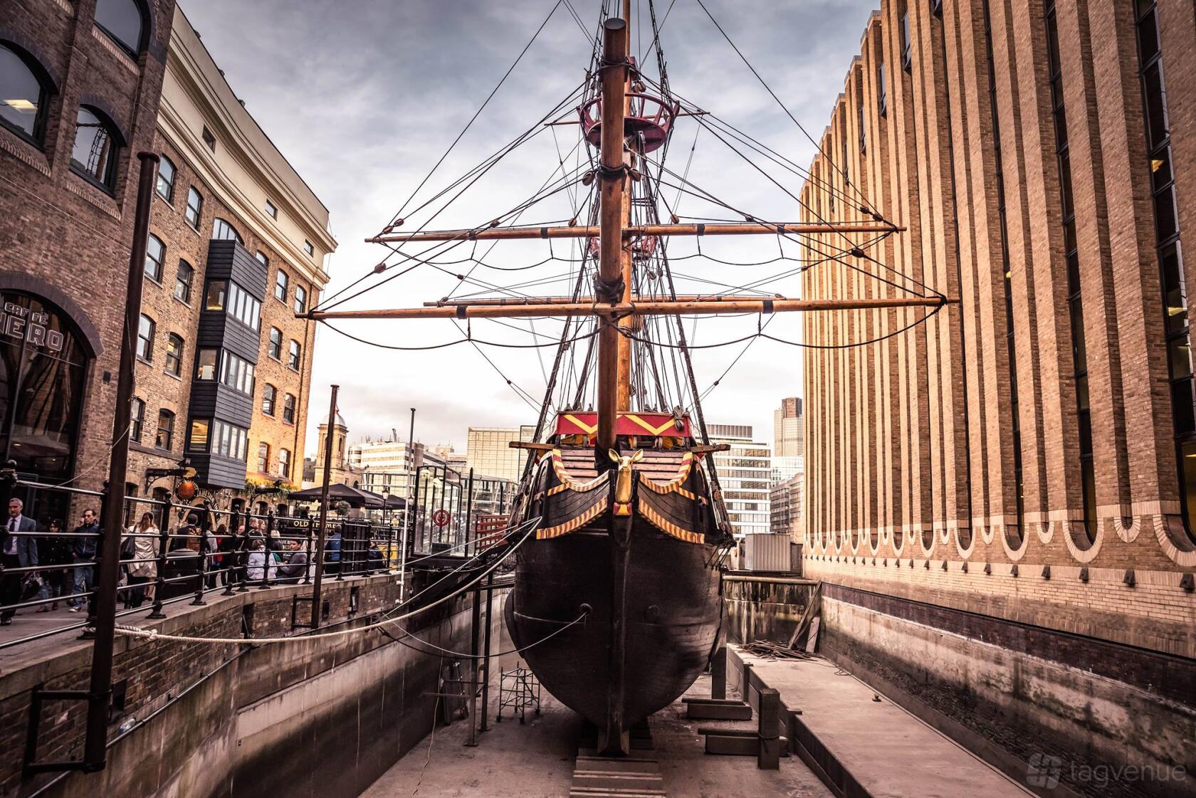 A historic ship museum docked between brick buildings with rigging, wooden hull, and city backdrop at The Golden Hinde.
