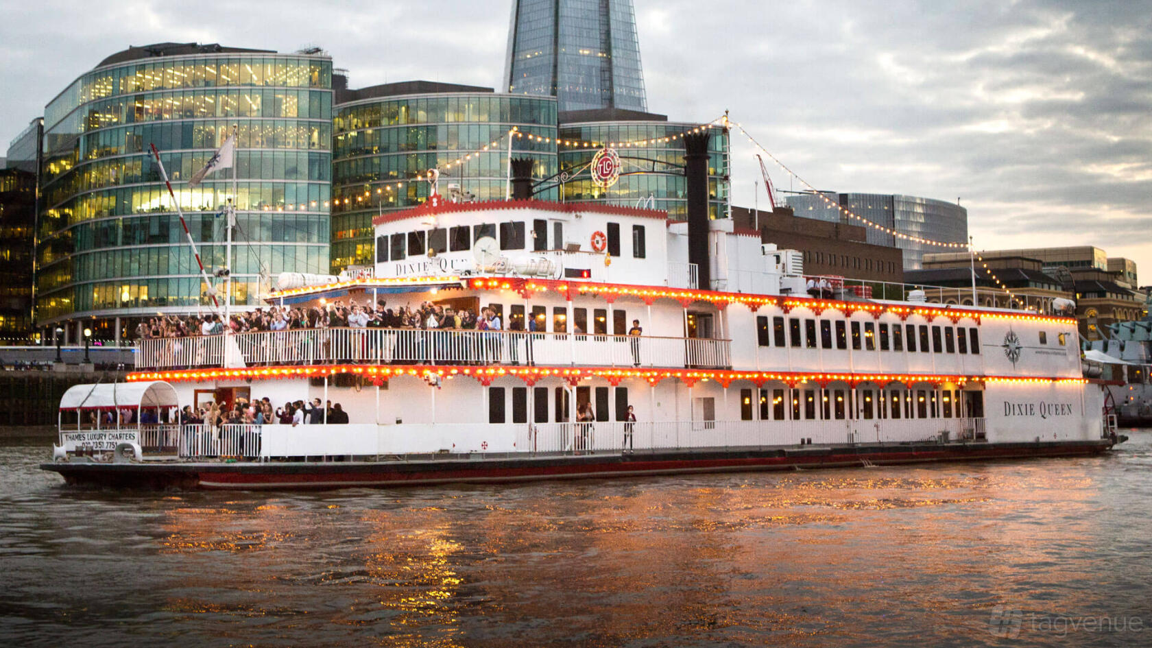 A large riverboat with festoon lights and open decks crowded with guests docked on the Thames at Dixie Queen – Thames Luxury Charters