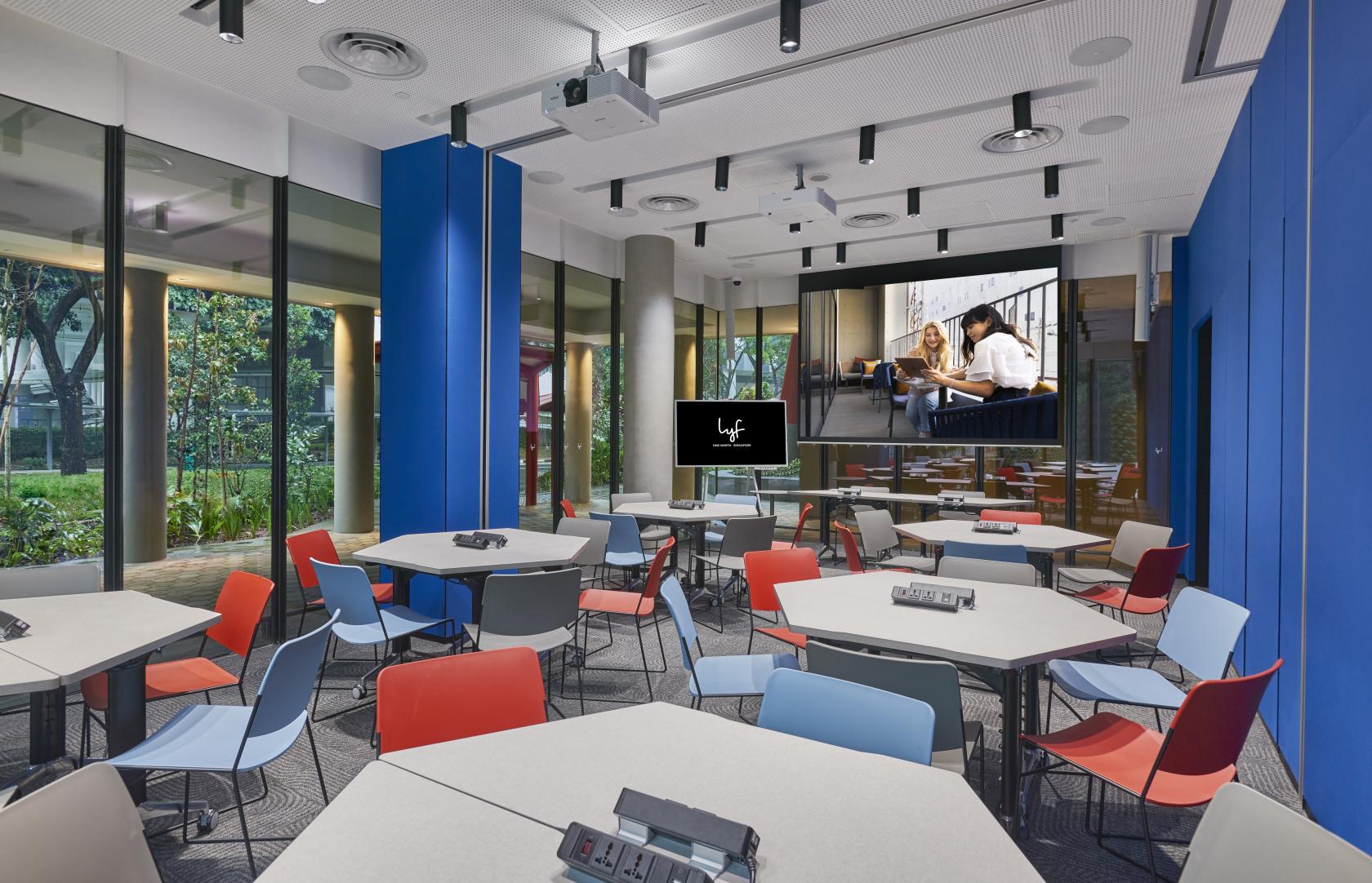 A meeting room with hexagonal tables, colorful chairs, floor-to-ceiling windows, and a projector screen at lyf one-north Singapore.