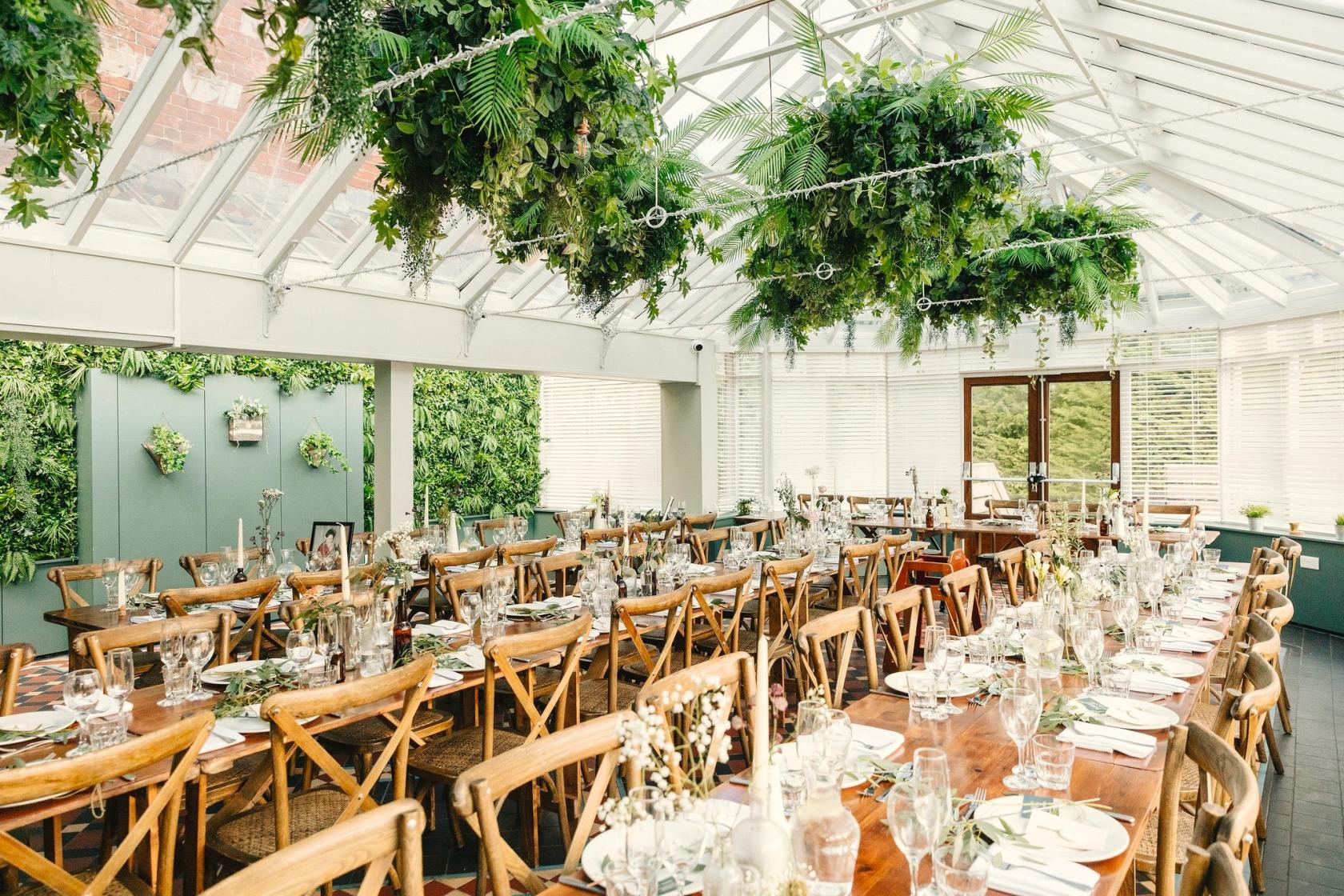 A conservatory with a glass ceiling, lush hanging greenery, and long wooden dining tables at The Faversham.