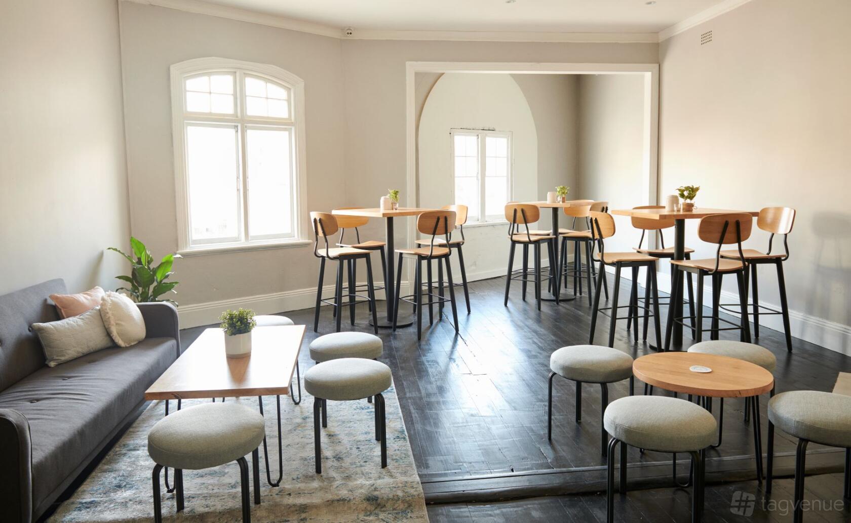 An upstairs lounge with wooden high tables, bar stools, a sofa, and large arched windows at Bondi Liquor Co.