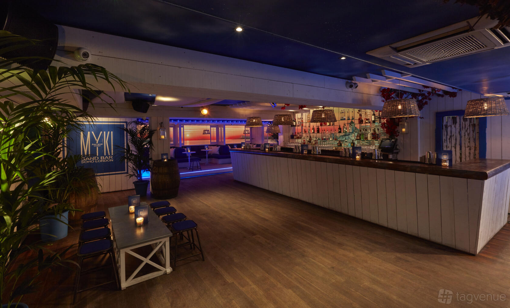 An event space in a nightclub with a long white bar, illuminated shelving, wooden floors, and lounge seating at Archer Street Soho.