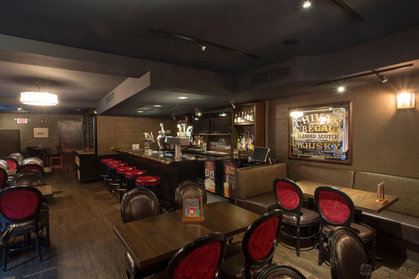 A pub interior with dark wood tables, red upholstered chairs, a long bar counter, and whisky signage at Duke of Kent.