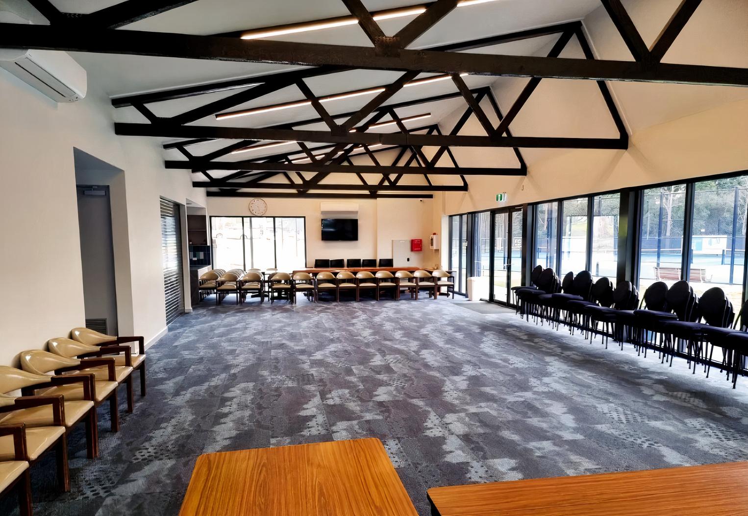 An event venue with exposed ceiling beams, carpeted floors, large windows, and stacked chairs at Frankston Centenary Tennis Club.