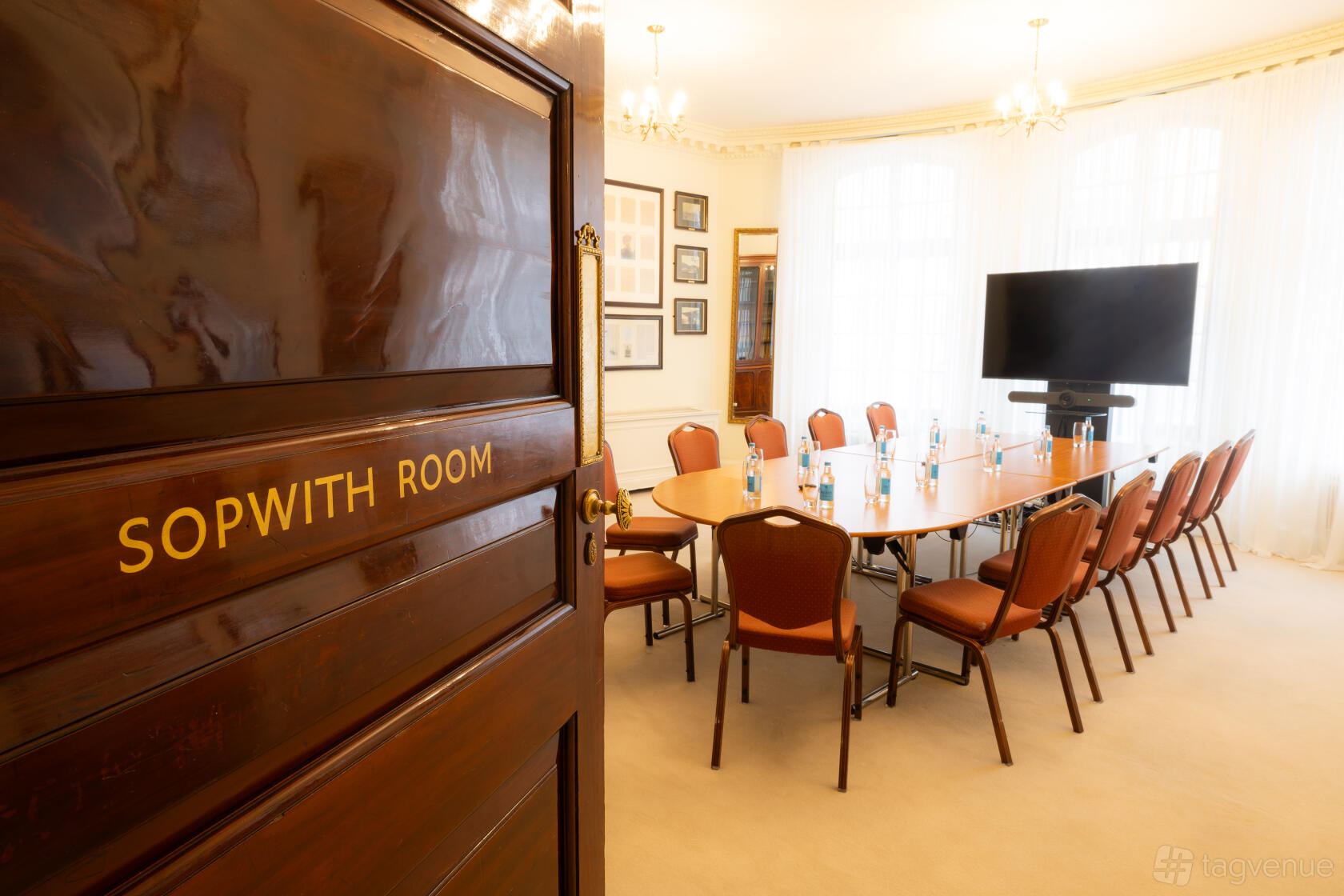 An event venue with a long oval table, brown chairs, large windows, and a TV screen in the Sopwith Room at No. 4 Hamilton Place.