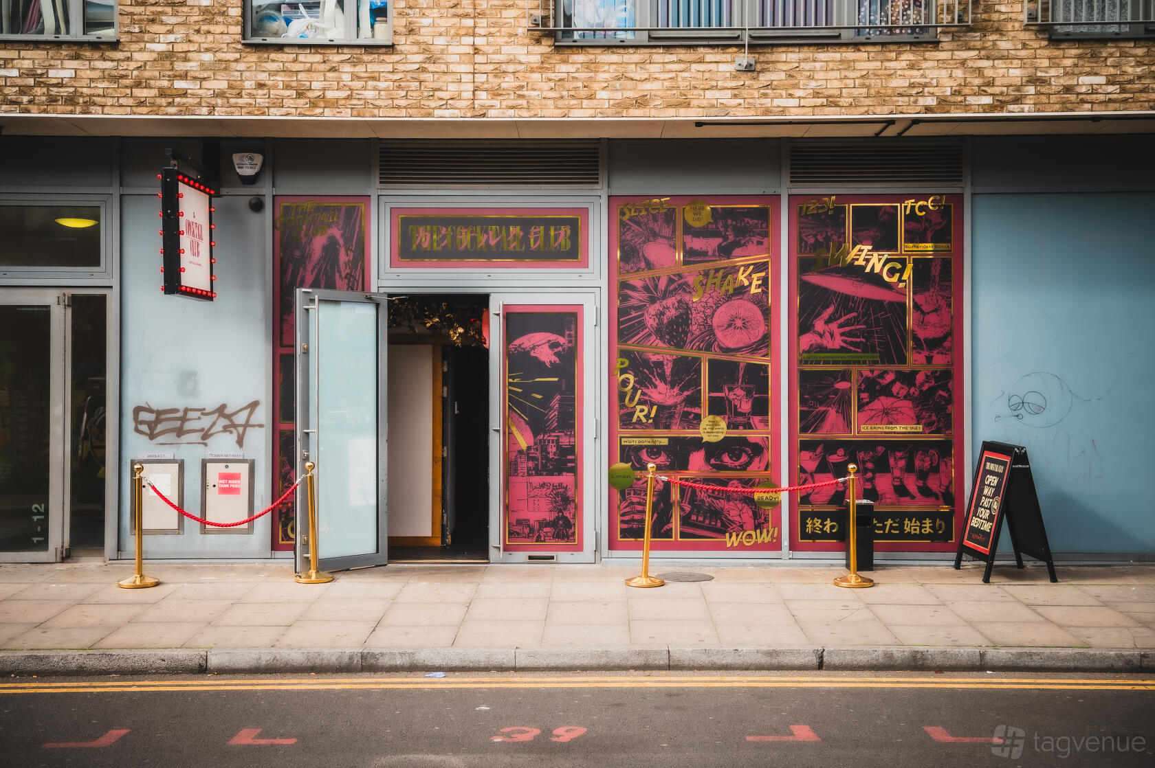 A pub with red and yellow graphic wall art, an open entrance door, and velvet ropes at The Cocktail Club Shoreditch.