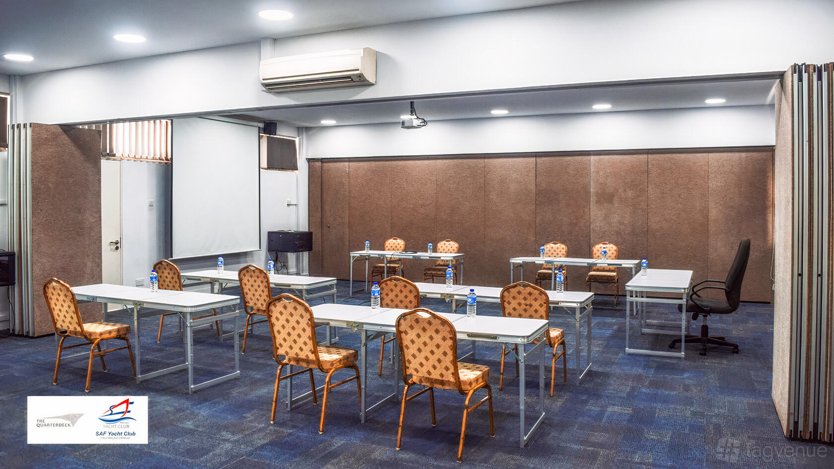 A seminar room with rows of tables, patterned chairs, a projector screen, and bottled water at SAF Yacht Club.
