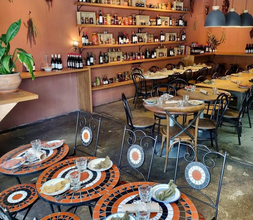 A restaurant with round mosaic tables, black chairs, and shelves displaying wine and pantry items at Pistou.