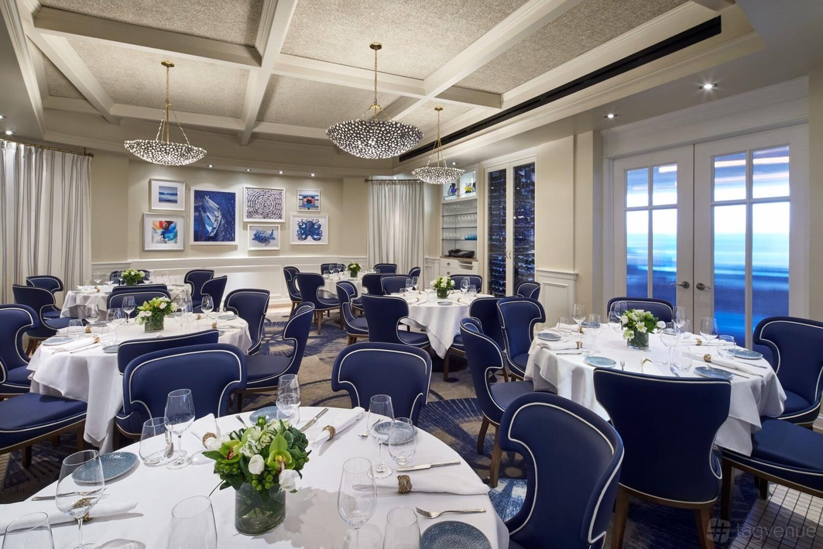 A function room with round tables covered in white linens, blue chairs, and chandeliers at Fiola Mare.