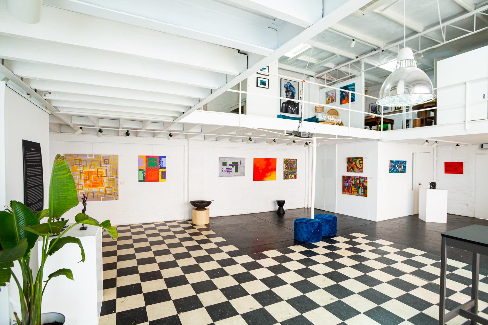A gallery with black and white checkered floors, white walls, colorful abstract art, and lofted ceilings at Artful Gallery.