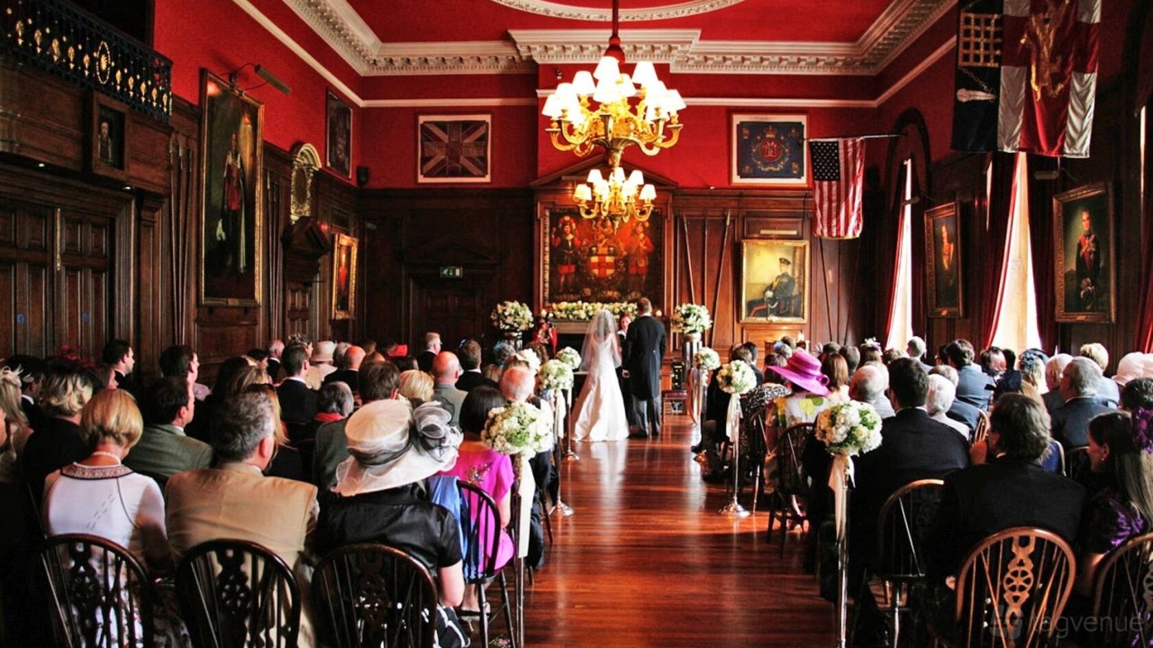 An event venue with ornate wood paneling, antique chandeliers, and red walls set up for a wedding ceremony at The HAC.