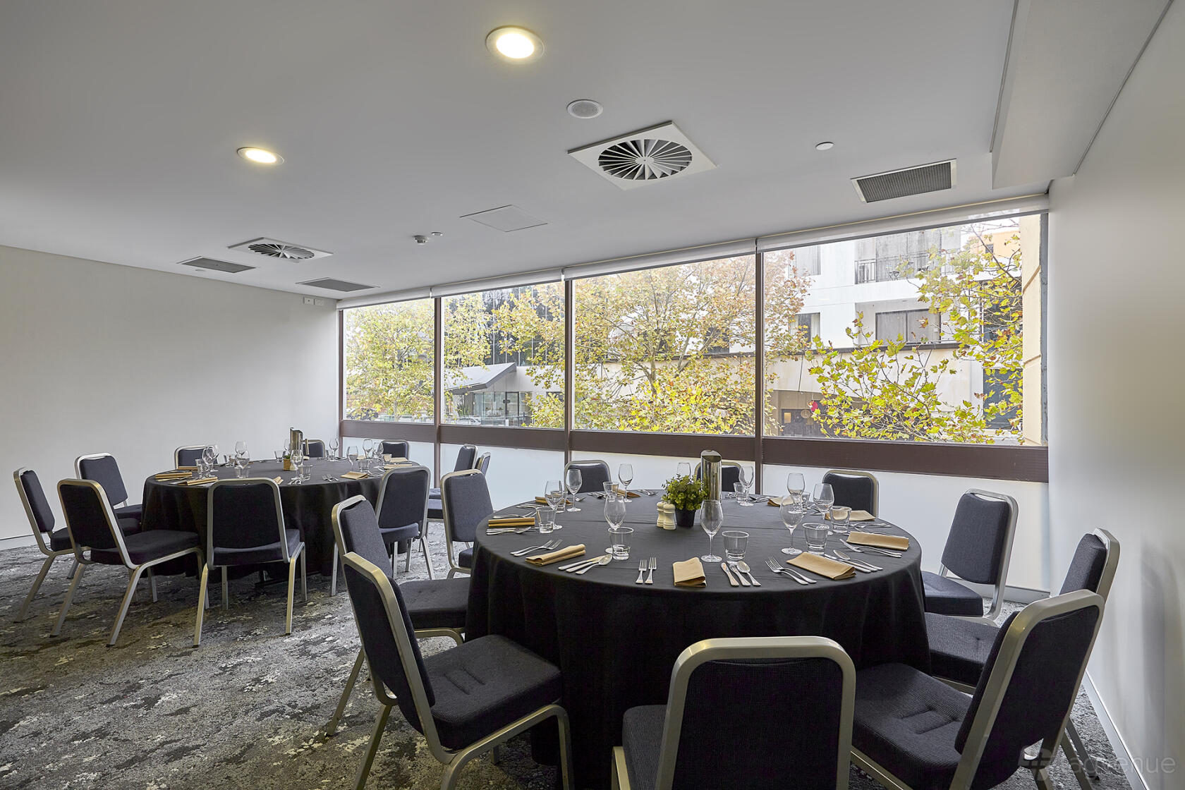 A hotel meeting room with floor-to-ceiling windows, round tables set for dining, and gray chairs at Mercure Perth on Hay.