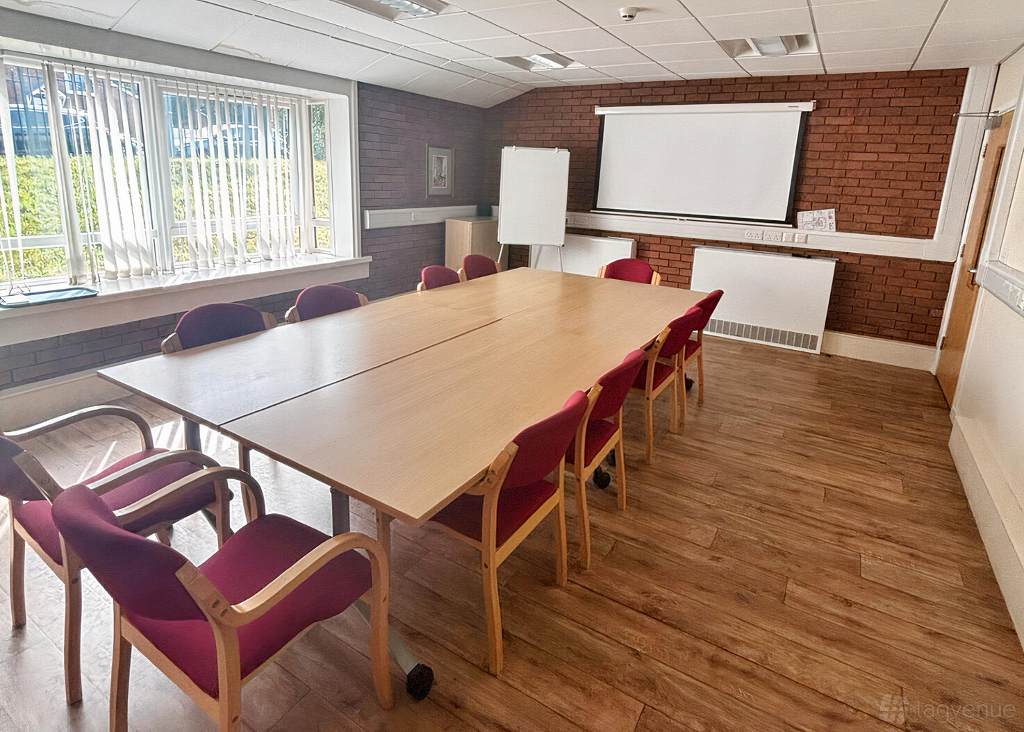 A meeting room with a large rectangular table, red cushioned chairs, wood flooring, and a projector screen at St. Sebastian's Community Centre.