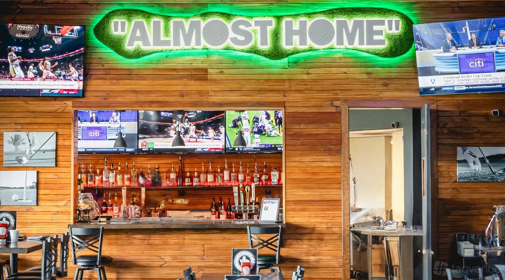 A restaurant bar with a wooden wall, neon 'Almost Home' sign, and multiple TVs above a liquor display at Three B’s on the Green.
