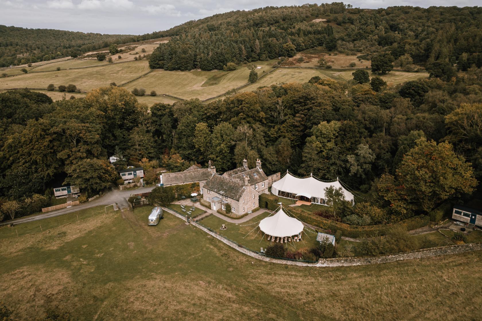 A country house with stone buildings, large tents, and rolling countryside surrounded by trees at Heathy Lea.