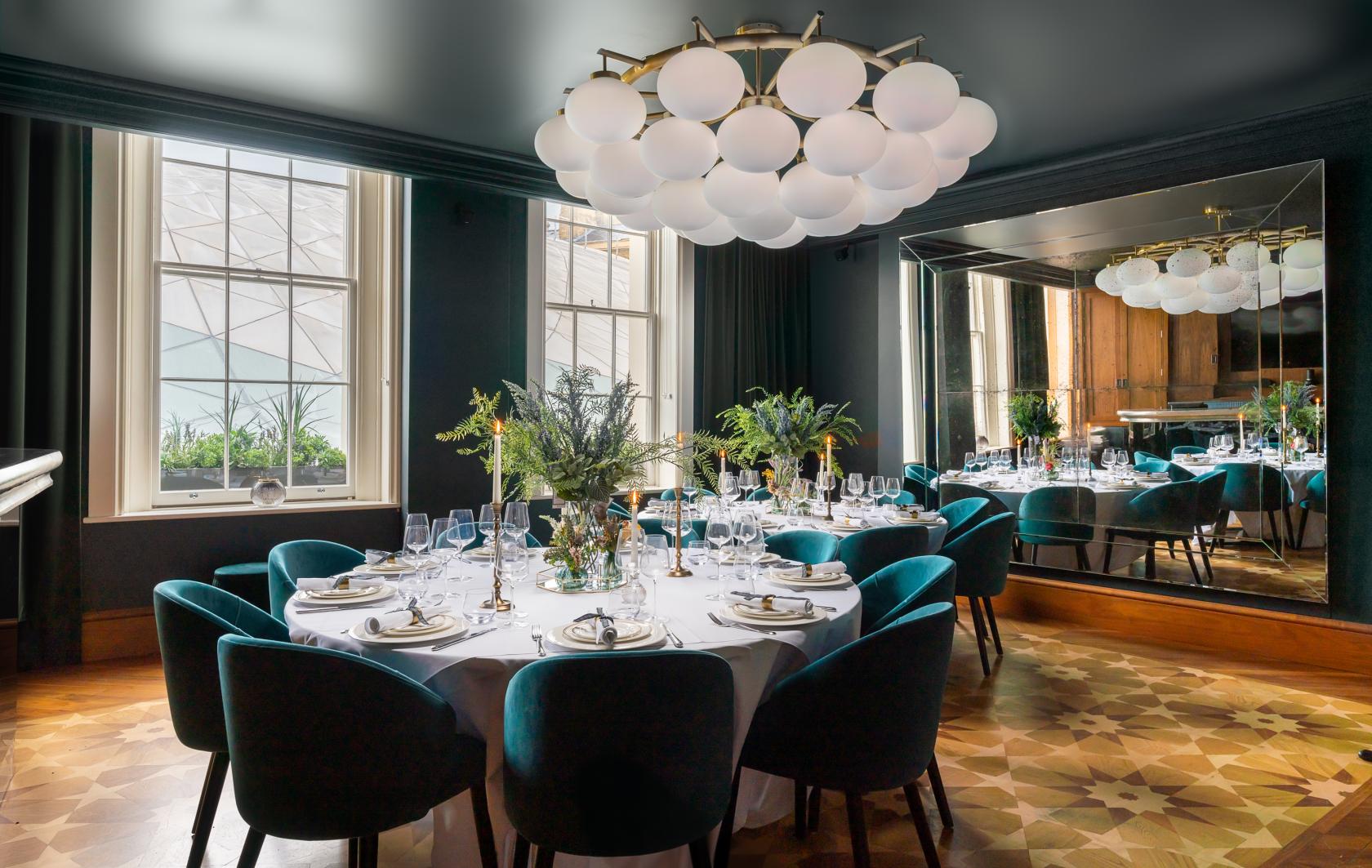 A hotel dining room with round tables, teal velvet chairs, a large modern chandelier, and a wall mirror at Great Northern Hotel: The Carriage & Lounge.