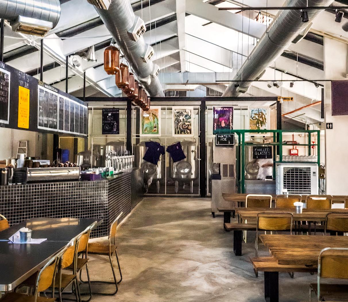 A pub with long wooden tables, metal chairs, and visible brewing tanks at Little Island Brewing Co. - Changi Village.