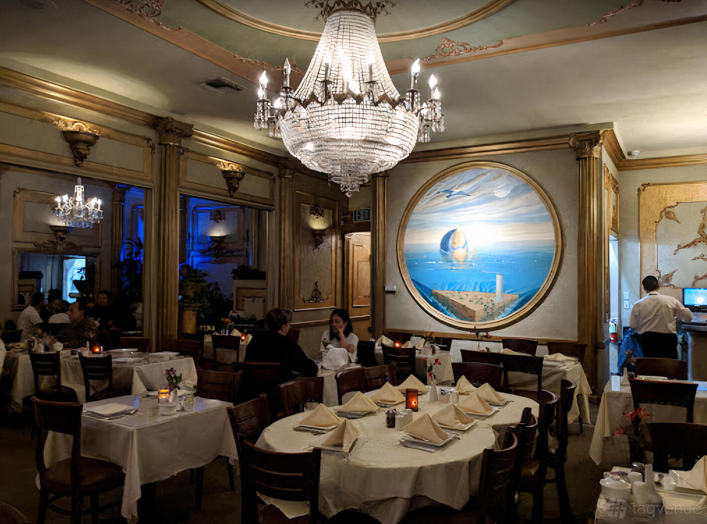 An event space in a restaurant with a grand crystal chandelier, round tables with folded napkins, and a mural at Darya Santa Monica.