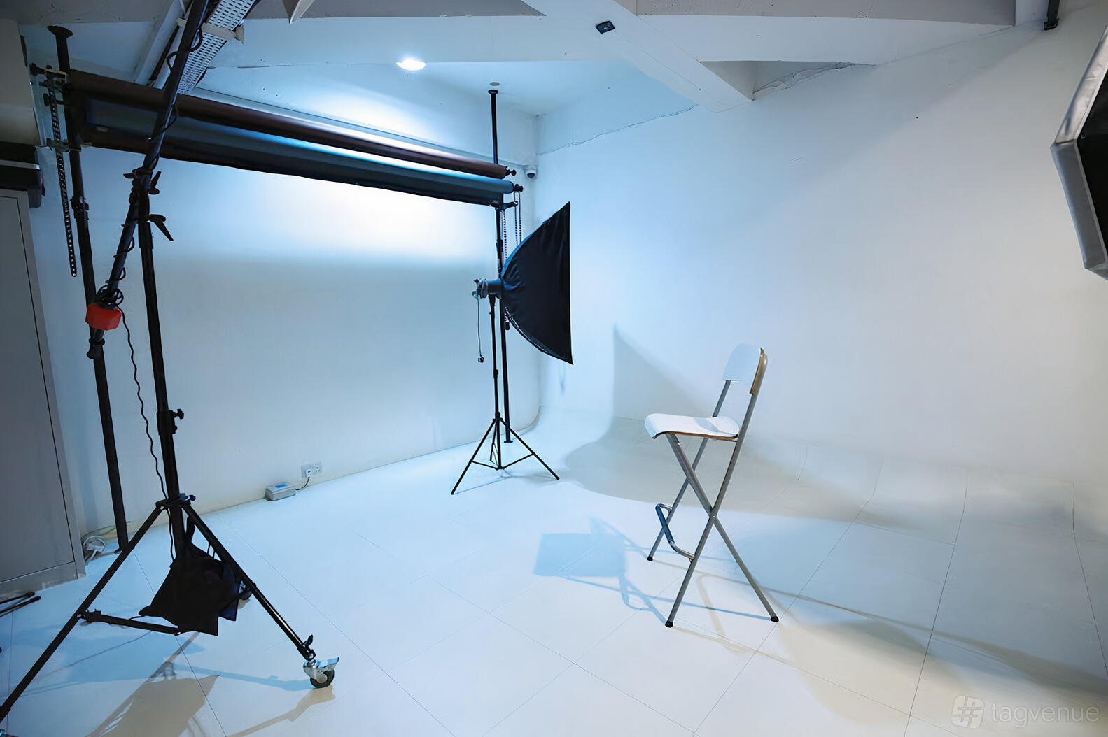 A photo studio with seamless white walls, a softbox light, backdrop system, and a folding chair at The Beauty Box Studio.