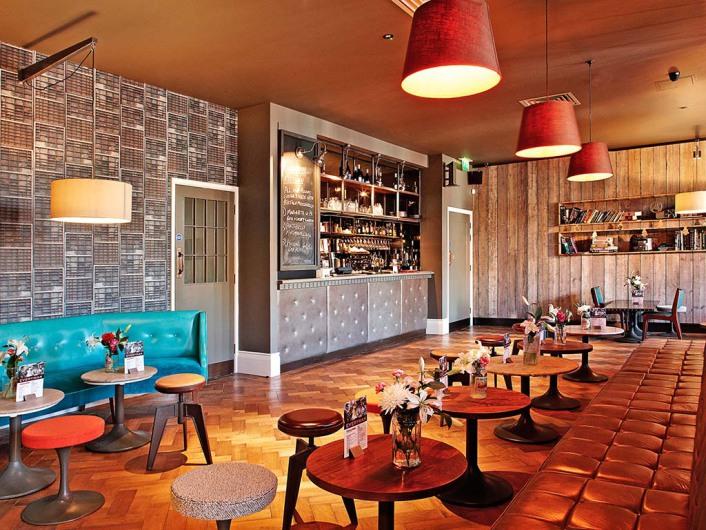 A cinema lounge with wooden floors, a teal sofa, round tables with flowers, and a bar at Everyman Maida Vale.
