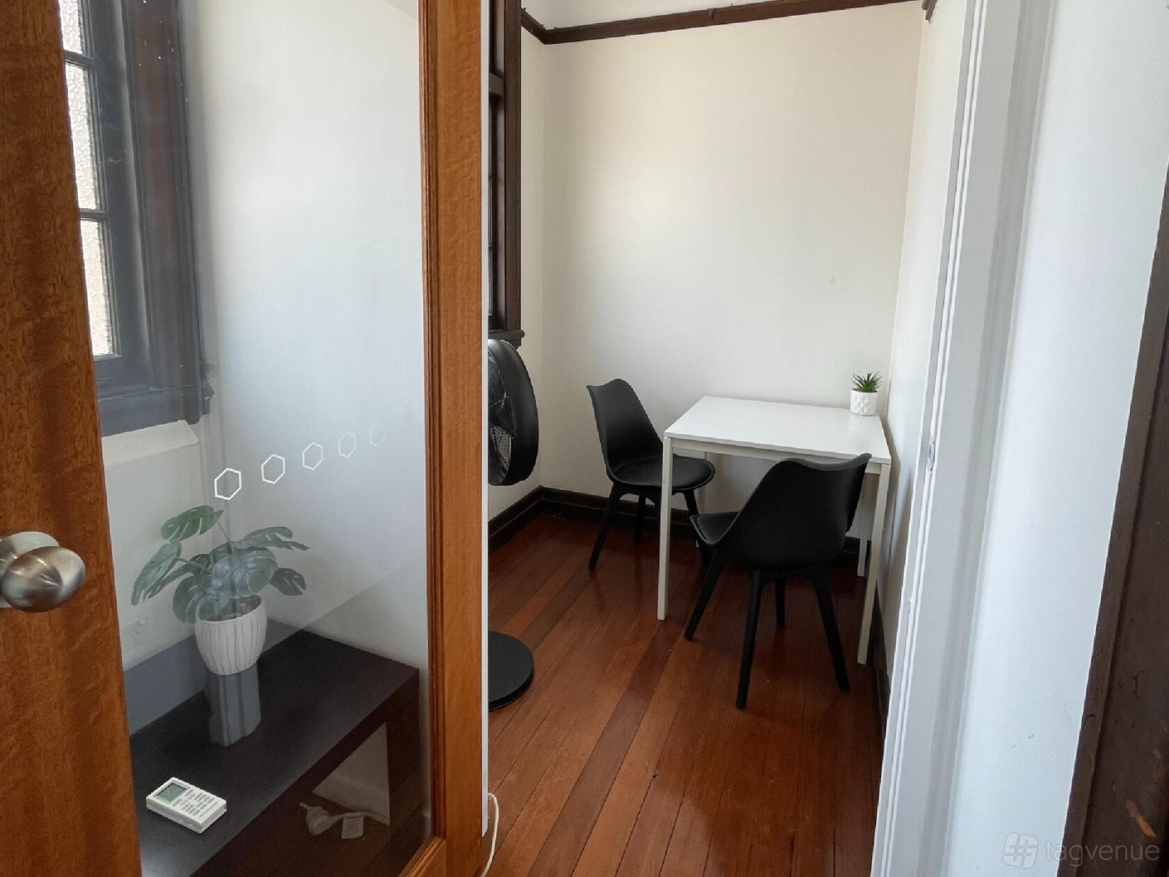 A compact meeting room with a small white table, two black chairs, wood flooring, and a potted plant at Salt Space Coworking - New Farm.