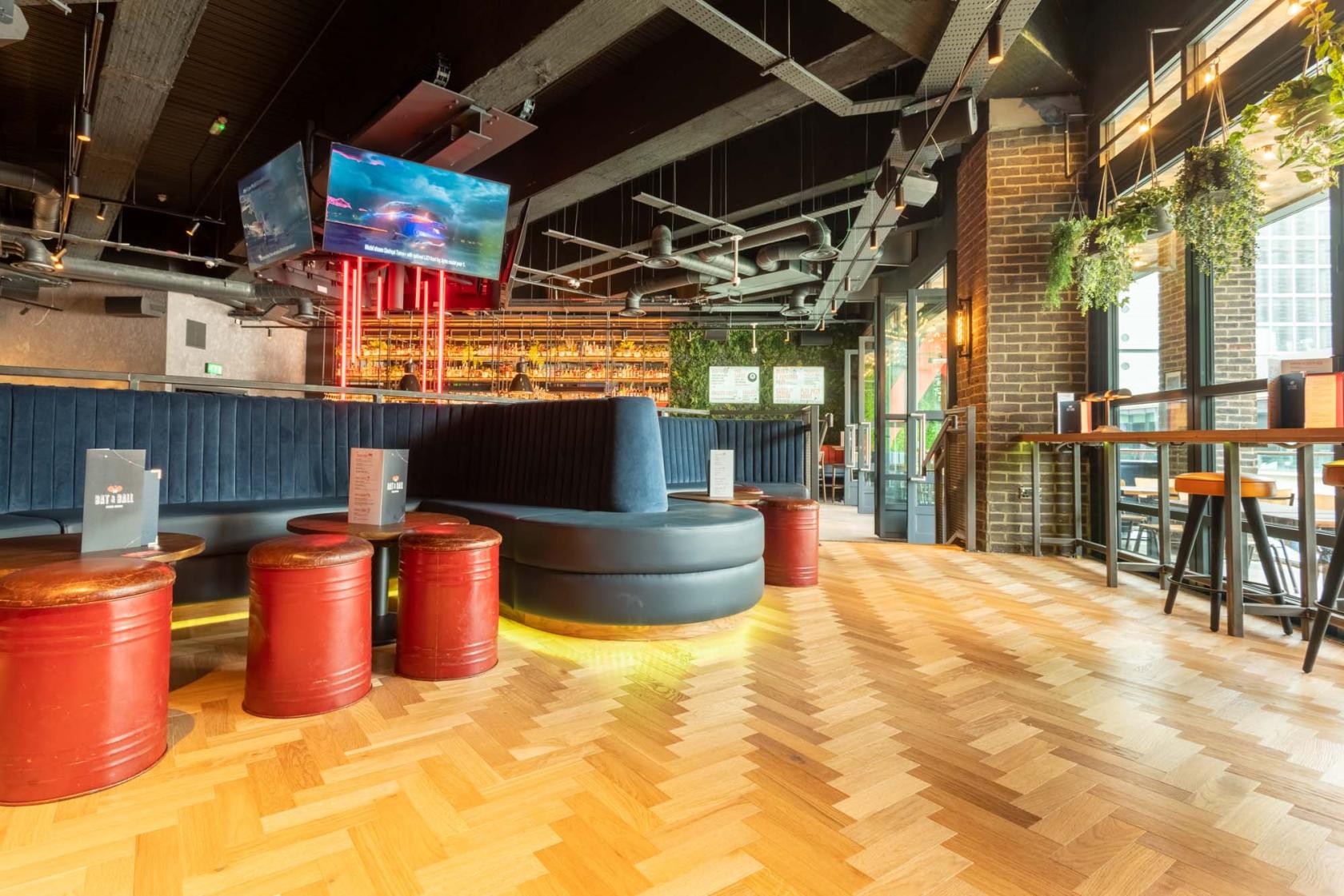 A lounge with curved booth seating, red drum stools, a large TV, and floor-to-ceiling windows at Bat & Ball.
