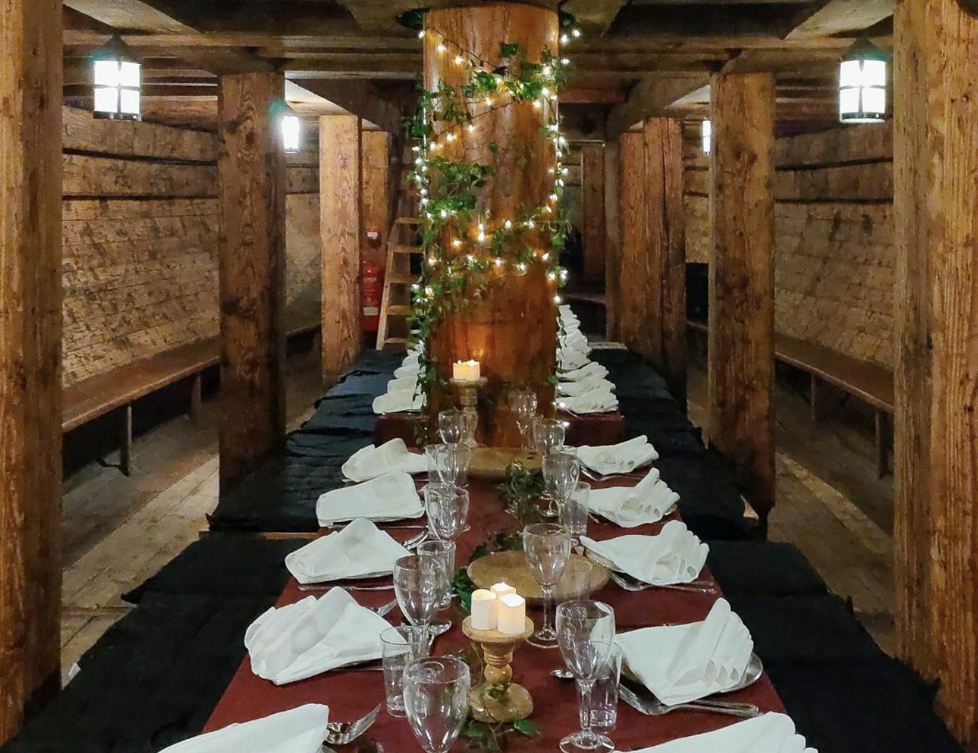 A historic ship museum with long wooden tables set for dining, surrounded by timber beams and string lights at The Golden Hinde.