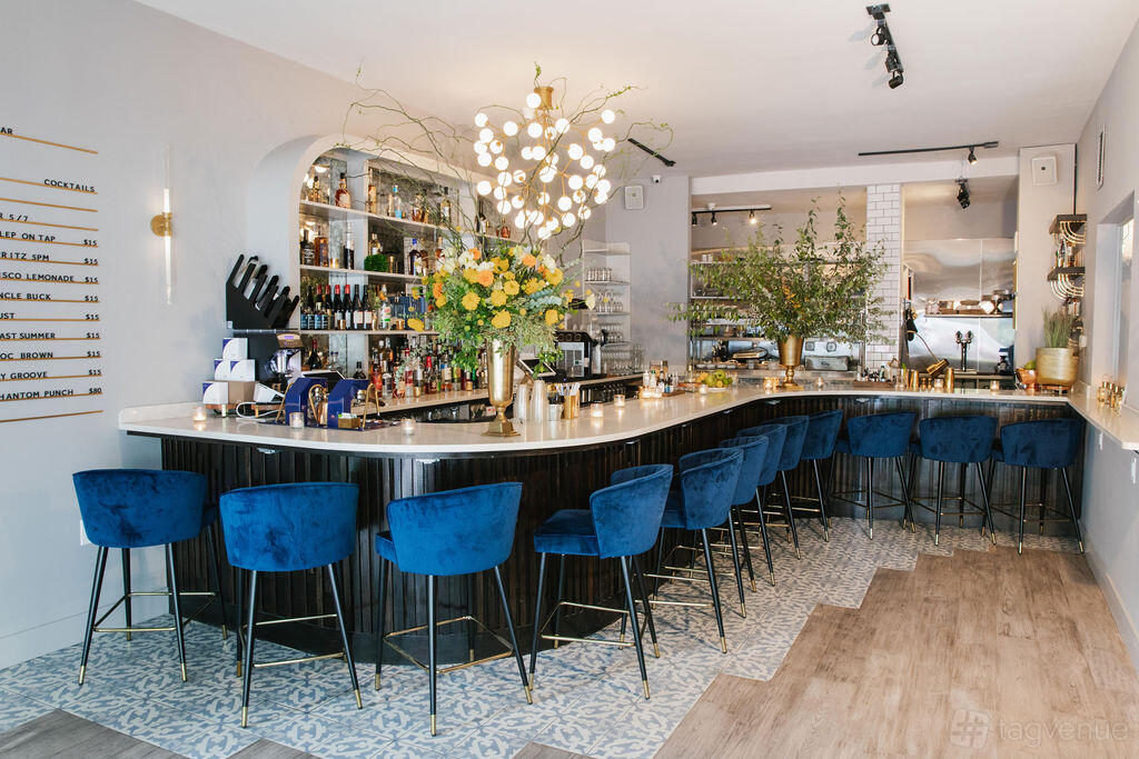 A restaurant with a curved bar, blue velvet barstools, a modern chandelier, and floral arrangements at Festival Cafe NYC.
