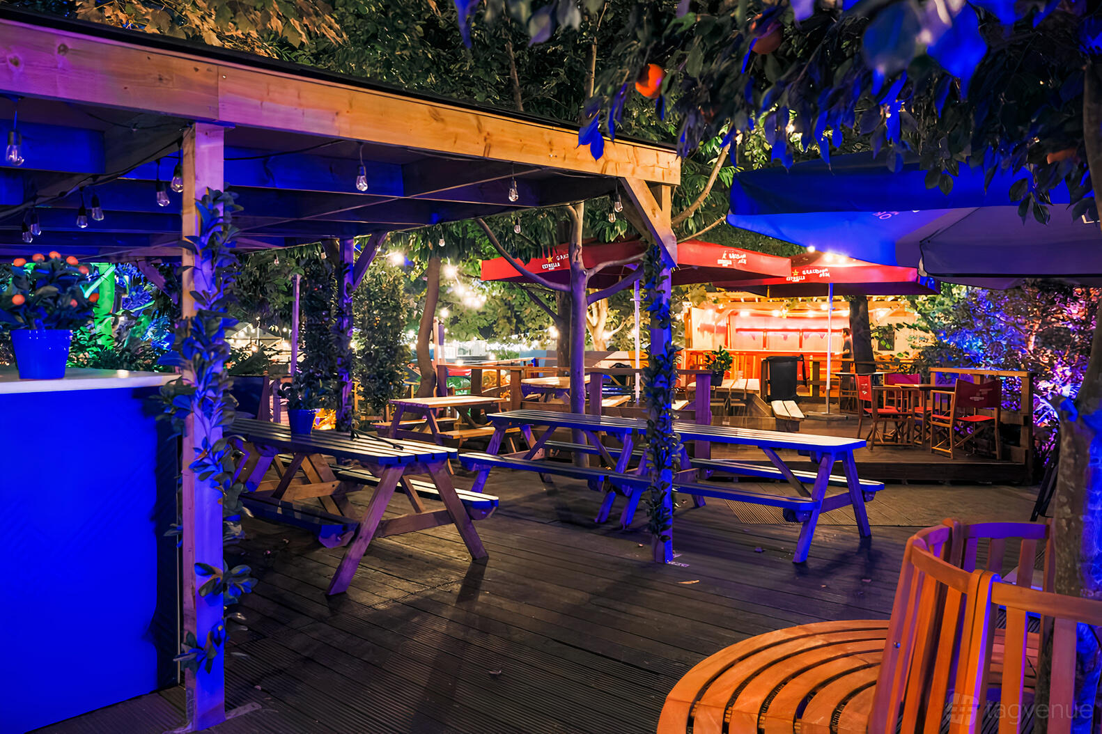An outdoor event venue with wooden picnic tables, deck flooring, and string lights under trees at Between the Bridges.