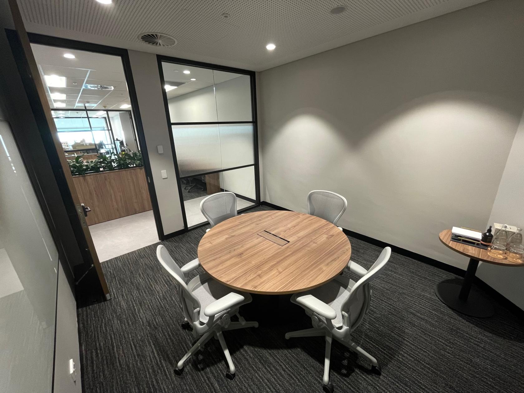 A meeting room with a round wooden table, four ergonomic chairs, and glass partition walls at Collins Place.
