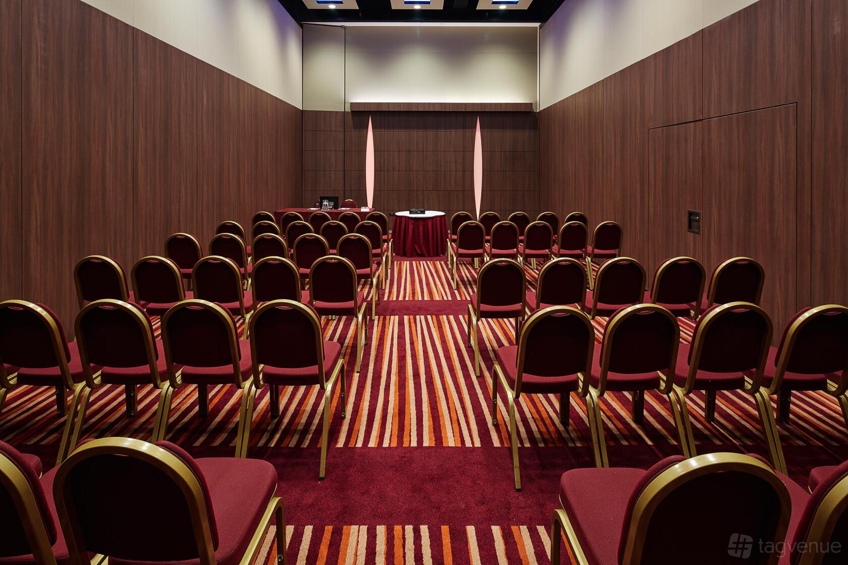 A meeting room with rows of red cushioned chairs, wood-paneled walls, and striped carpeting at Mercure London Earl's Court.