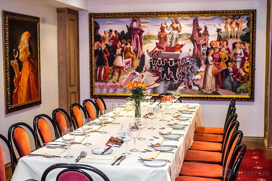 A dining room with a long table set with glassware, red chairs, and colorful wall art at Bolton's Venetian Room.