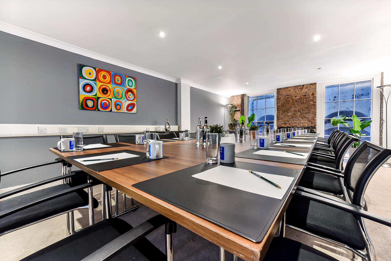 A meeting room with a long conference table, black chairs, exposed brick wall, and colorful wall art at Adam House Event Space.