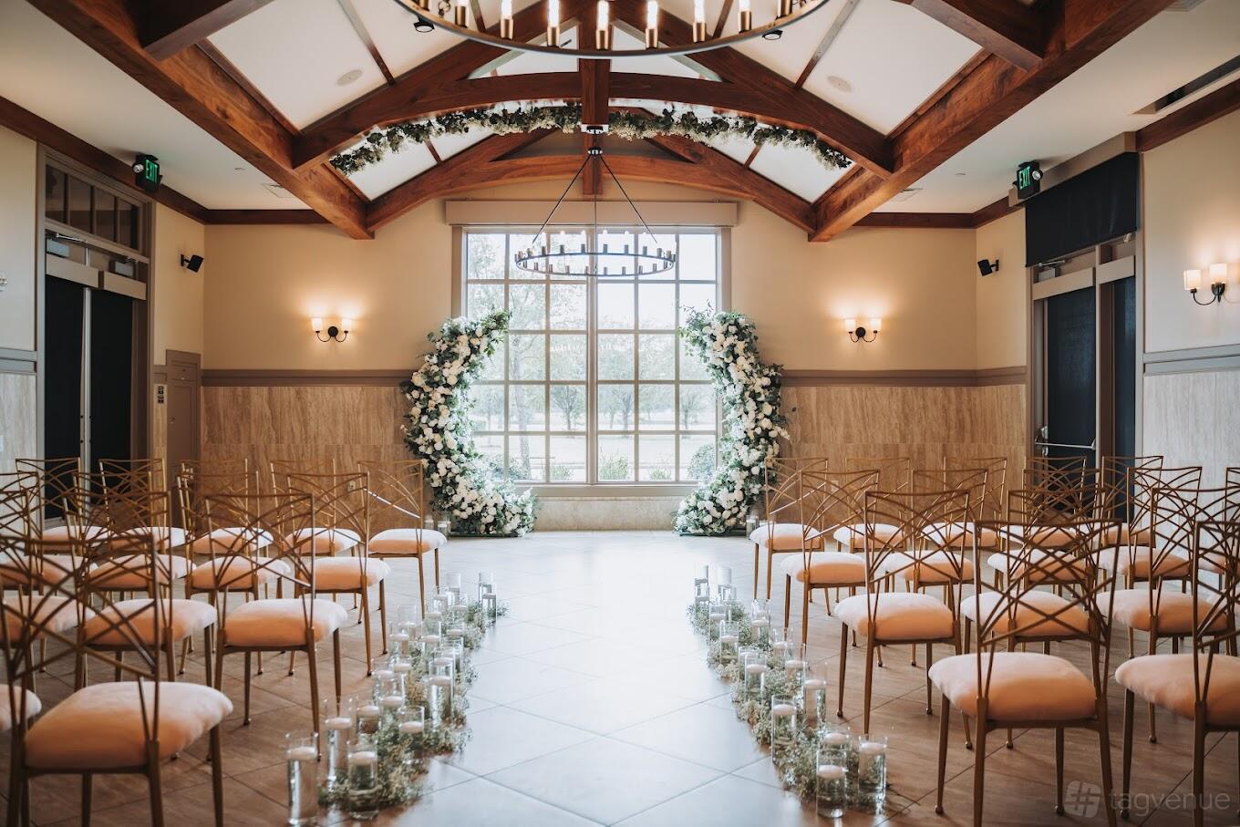 A dining room with arched wooden beams, a large window, floral arch decor, and rows of gold chairs at Milano Event Center.
