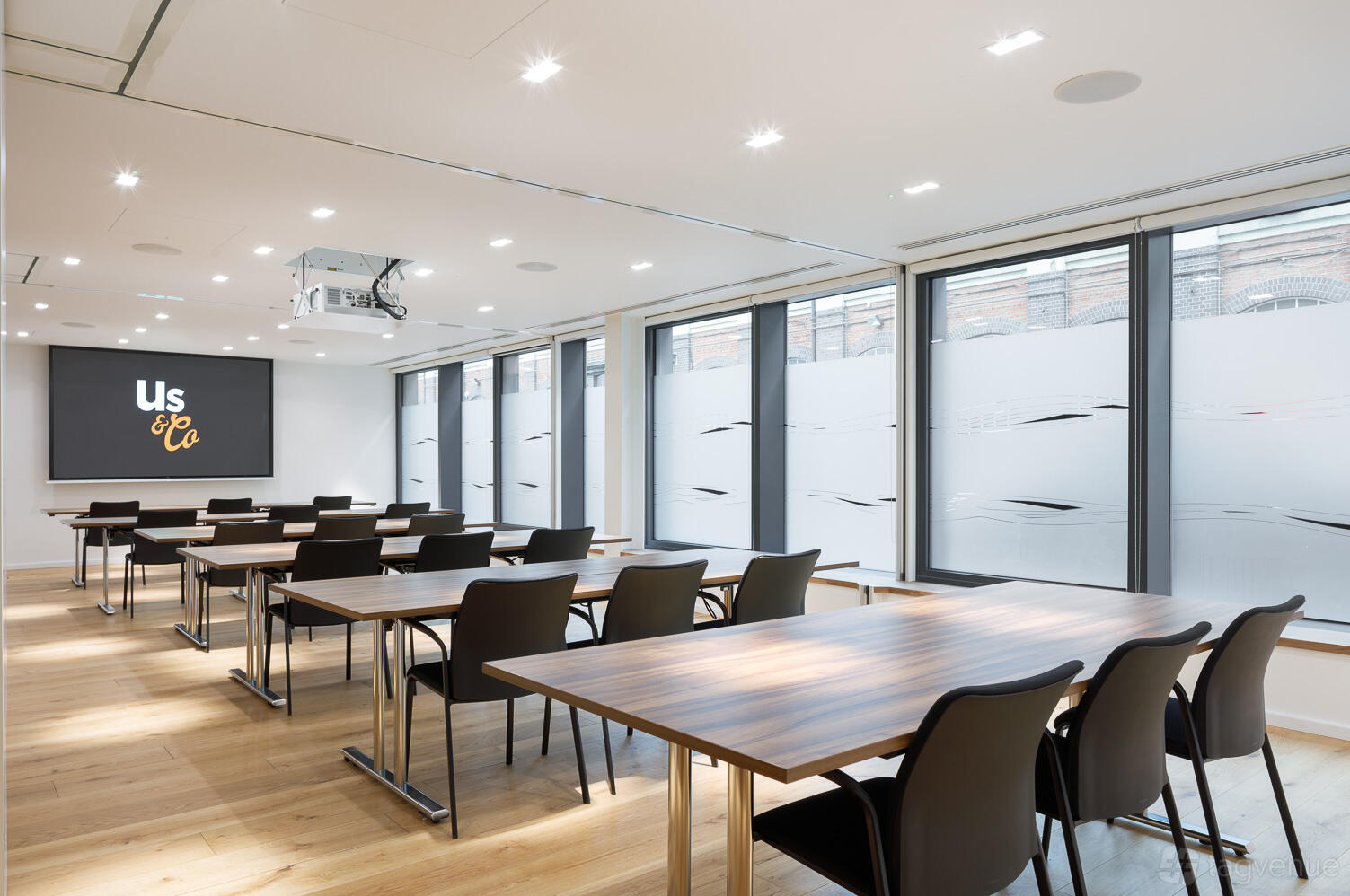 A conference centre with rows of wooden tables, black chairs, large windows with frosted glass, and a projector at Us&Co Stratford.