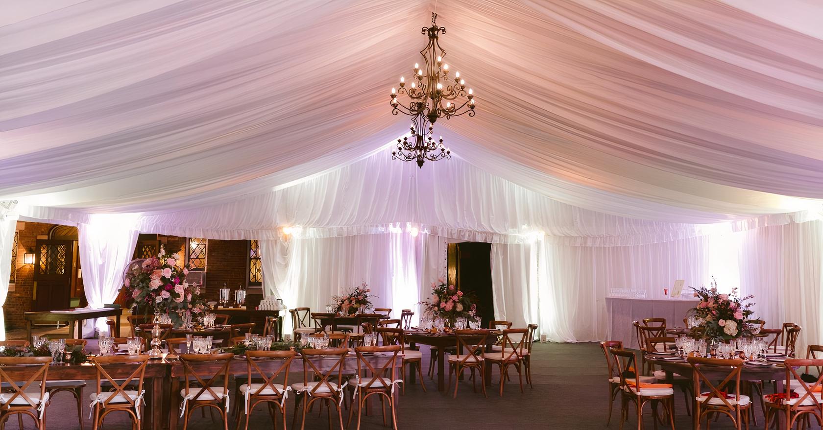 An event space with draped white ceilings, chandeliers, and wooden tables with floral centerpieces at Lyndhurst Mansion